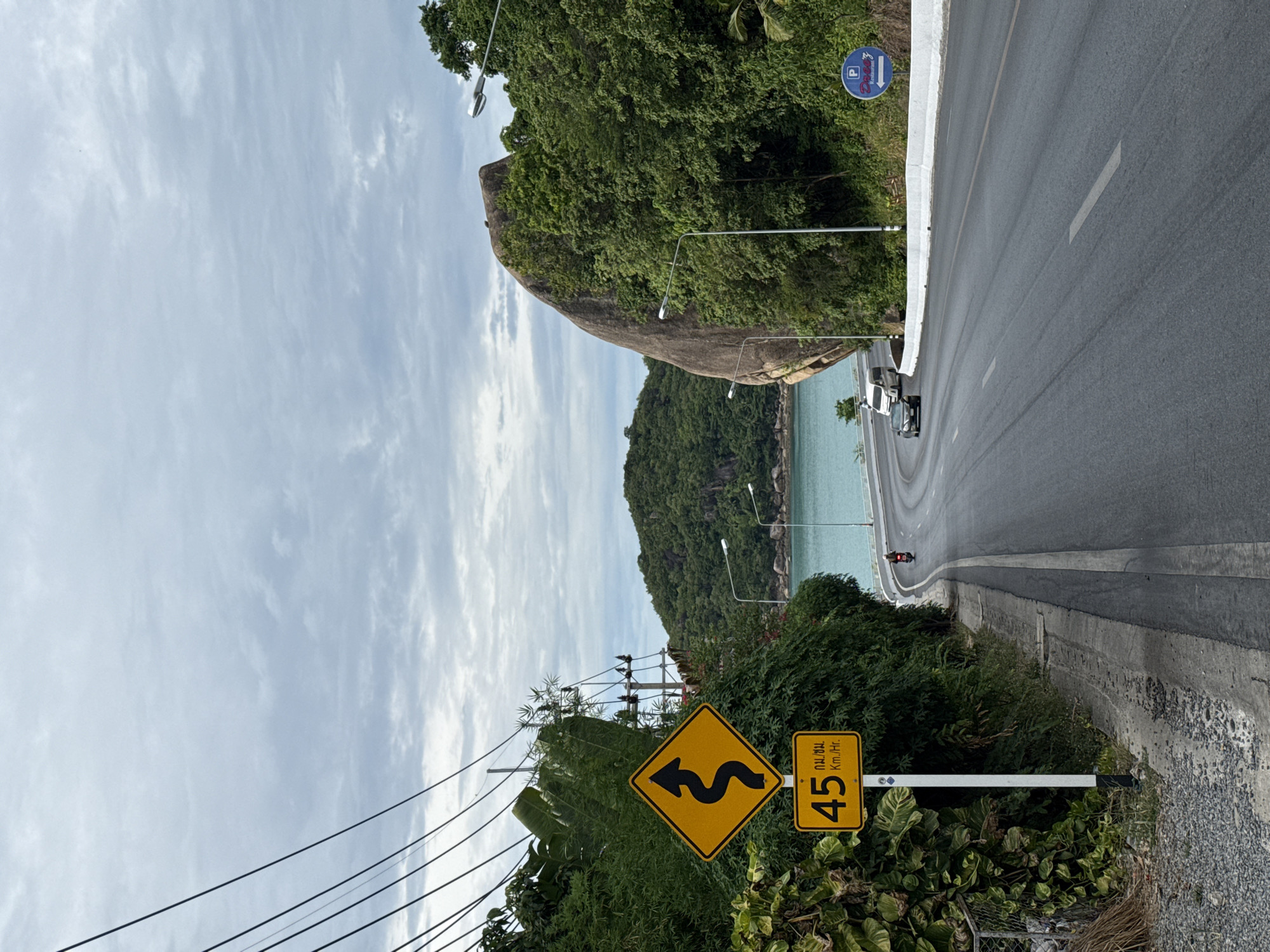 This is a photograph of a winding road on the island of Ko Samui, Thailand. The road curves to the right, with the turquoise sea visible on the left, bordered by lush green vegetation and a large rock formation. Overhead power lines are strung across the sky. In the foreground, a yellow diamond-shaped sign with a black zigzag arrow indicates a winding road, with a smaller rectangular sign below it displaying "45 Km./Hr.". Further down the road, a white van and a black car are visible, along with a person riding a motorbike in the distance. A blue parking sign with a white "P" and an arrow pointing to the left is mounted on a pole to the right of the road. The sky is overcast with grey clouds.