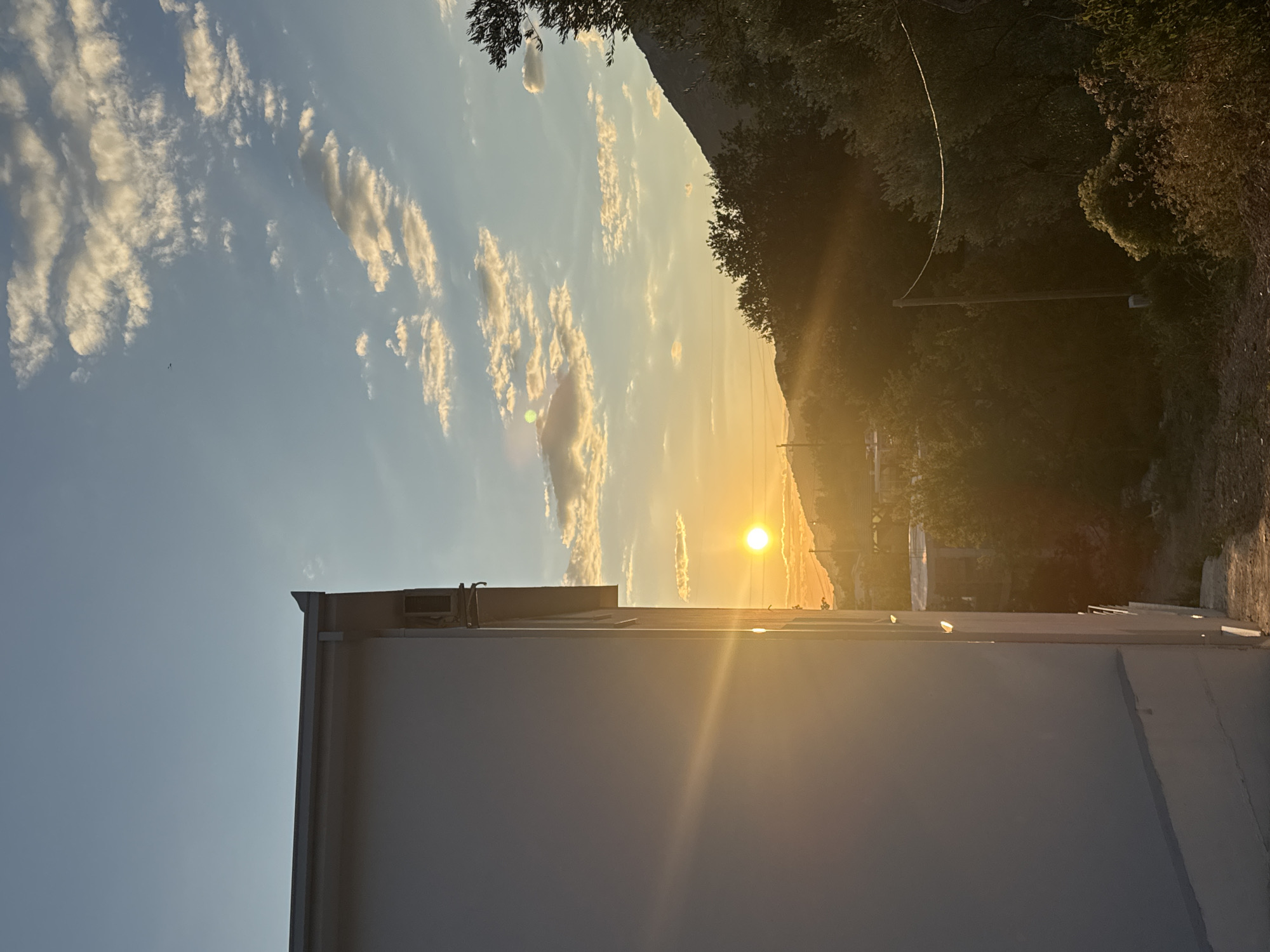 The image depicts a serene sunset scene in Sustaš, Montenegro, viewed from the side of a white building. The sun, a bright orb, is positioned low on the horizon, casting a warm golden glow across the landscape. Rays of light emanate from the sun, illuminating the sky and the surrounding vegetation. Wispy clouds are scattered across the clear blue sky, tinged with the colors of the sunset.
In the foreground, the stark white wall of a building occupies the left side of the frame. An air conditioning unit is visible mounted on the exterior wall near the top edge. The building's roofline is also visible, with a gutter running along its edge.
The middle ground features a dense expanse of lush green trees and foliage, with the sun's rays filtering through them. A wooden utility pole stands amidst the trees, with a power line stretching across it. In the background, rolling hills and mountains are silhouetted against the vibrant sky. The overall impression is one of tranquility and natural beauty. The image was taken in Sustaš, a locality within the municipality of Bar, Montenegro.