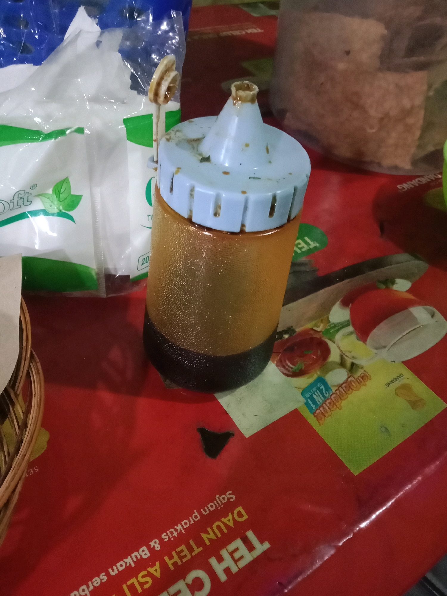 A close-up, eye-level shot captures a condiment bottle and a tissue pack on a red table, likely in Semarang, Indonesia, given the visible text. The condiment bottle, made of translucent orange plastic with a white cap, is filled with a dark brown liquid, possibly soy sauce or a similar condiment. It appears slightly used, with some residue on the cap and neck. To the left of the bottle, a partially opened, clear plastic-wrapped tissue pack is visible, featuring green leaf graphics and text. The red table surface is cluttered with various items, including small packets with colorful graphics, and a wicker basket edge on the left. In the background, out of focus, is a large, opaque container filled with a light brown granular substance, and a blurred, brown patterned surface. The lighting is dim, creating shadows and highlighting the textures of the objects.