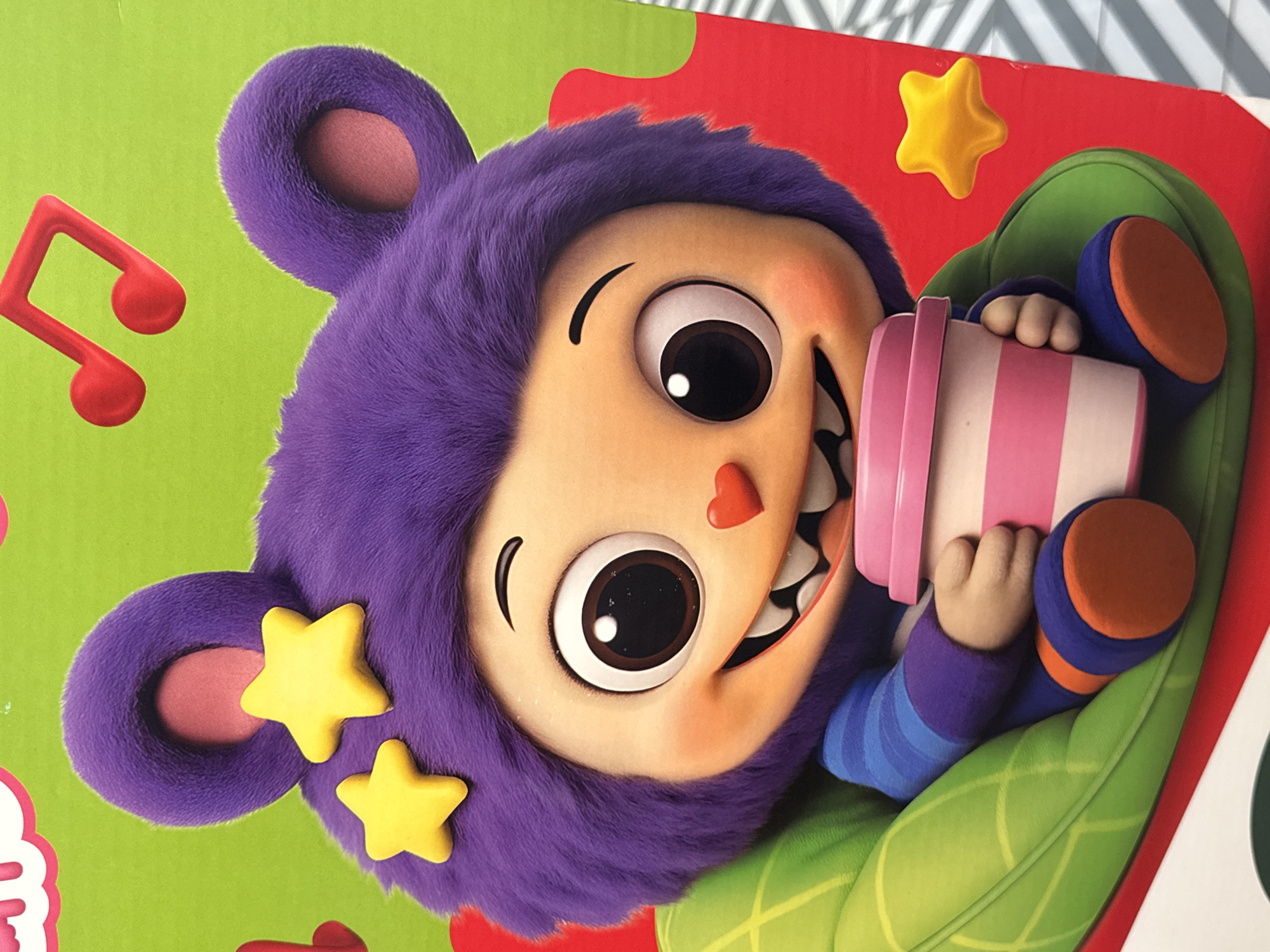 A character, possibly a cartoon, with purple furry ears and hair is depicted. The character has large eyes, a smiling mouth with sharp teeth, and a heart-shaped nose. It is wearing a blue and purple striped shirt and blue pants with orange accents. The character is sitting in a green, leaf-shaped chair and holding a pink and white striped cup. Yellow stars are visible on the character's head and on the red background. Musical notes are also present on the green background. The image appears to be a product advertisement or illustration. There is no information within the image that indicates a location in Mikhaylovsk, Russia.
