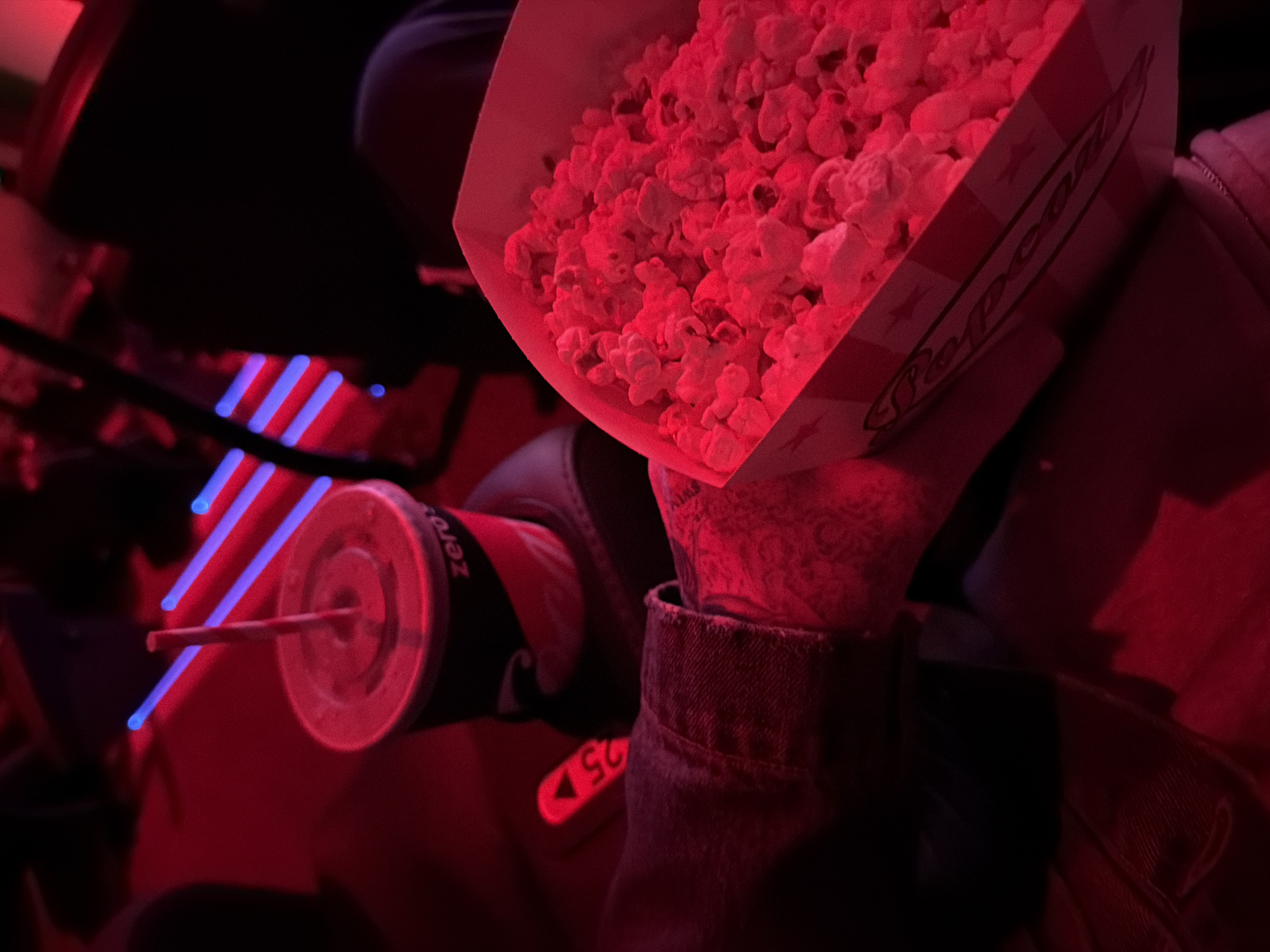 The scene depicts a person sitting in a movie theater, illuminated by a red light. They are holding a box of popcorn in their right hand and a drink labeled "Zero" in the left-hand side of the image. The seat number "25" is visible on the armrest. There are blue light stripes, likely from the theater's ambiance, on the left. The setting and objects suggest a movie theater. This location is not specific to the city of London, United Kingdom.