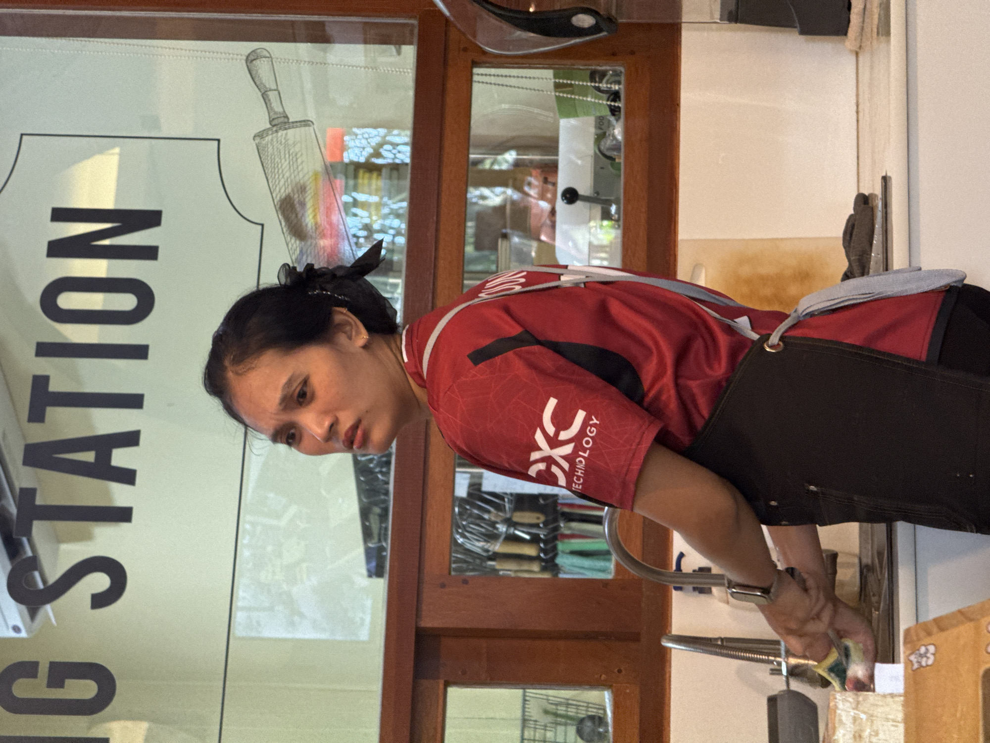 In the image, a woman is shown in a kitchen area, wearing a red shirt with black accents and a black apron. She is standing at a sink and appears to be washing dishes, holding a sponge in her right hand. Behind her, a window reveals reflections of the outdoor environment and some kitchen utensils are visible in racks on the wall. A sign above her head reads "IG STATION". The lighting suggests an indoor setting with natural light coming from the window. The overall scene depicts a working environment, possibly a restaurant or cafe, in ตำบลเกาะพะงัน, Thailand.