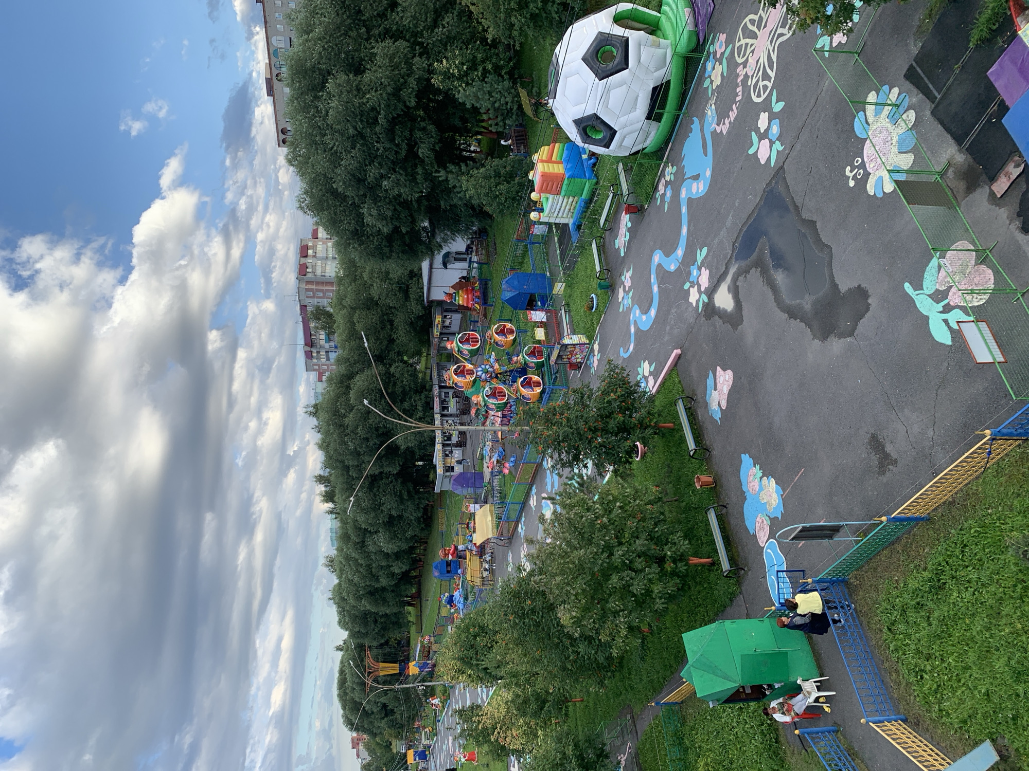 The image depicts an outdoor amusement park, possibly a playground or a small carnival, located in Prokopyevsk, Russia. The park features various attractions including a Ferris wheel with multiple colorful gondolas, an inflatable soccer ball structure, and an inflatable bouncing castle. The ground is paved with asphalt and decorated with painted designs of flowers and a blue wavy line, suggesting it is intended for children.
Several people are present in the park. In the foreground, two individuals stand near a green structure, one in a yellow jacket and the other in a dark coat. Further to the left, two women are seated on chairs, one appearing to be holding a phone. Benches are scattered throughout the park, and trash receptacles are visible.
The park is bordered by a fence with yellow and blue bars. Lush green trees form a dense backdrop, with apartment buildings visible in the distance, characteristic of urban Russian architecture. The sky is partly cloudy, with patches of blue sky visible amongst dramatic, gray clouds. The overall atmosphere suggests an overcast day, possibly after rain, given the puddles on the asphalt. Streetlights with distinctive curved arms are positioned along a pathway.