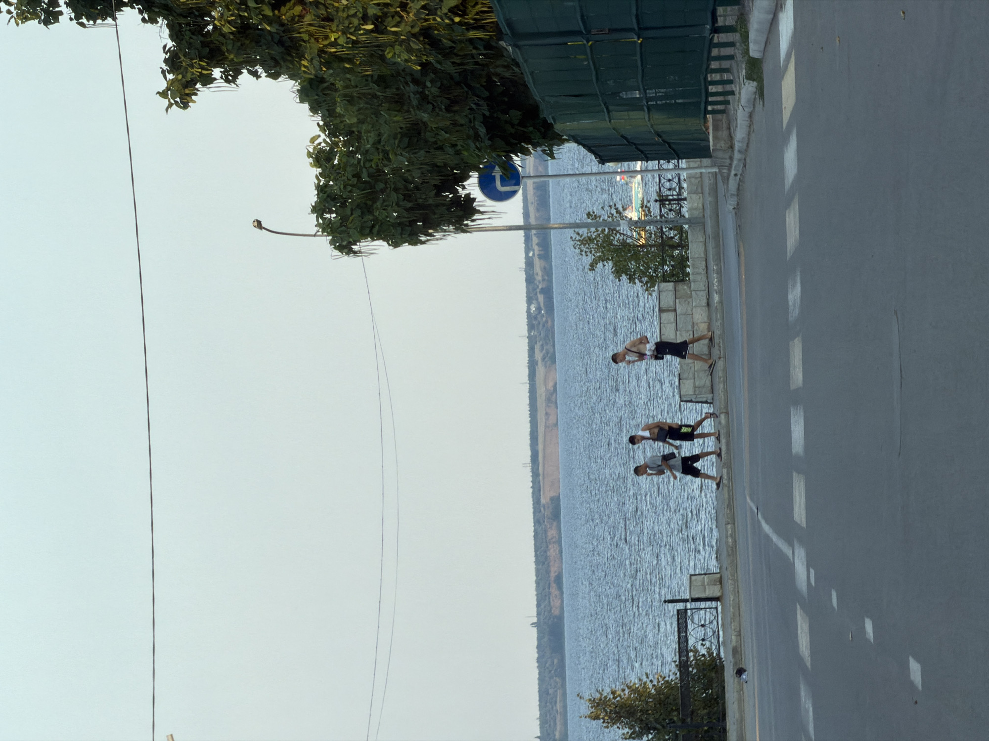 The image shows a street with a crosswalk in the foreground and a body of water in the background. Three young men are walking on a sidewalk next to the water. There are trees and a fence along the sidewalk. A street lamp is visible on the right side of the image. The sky is clear and the water appears calm. The location is likely a waterfront area in Kamyshin, Russia, given the context.