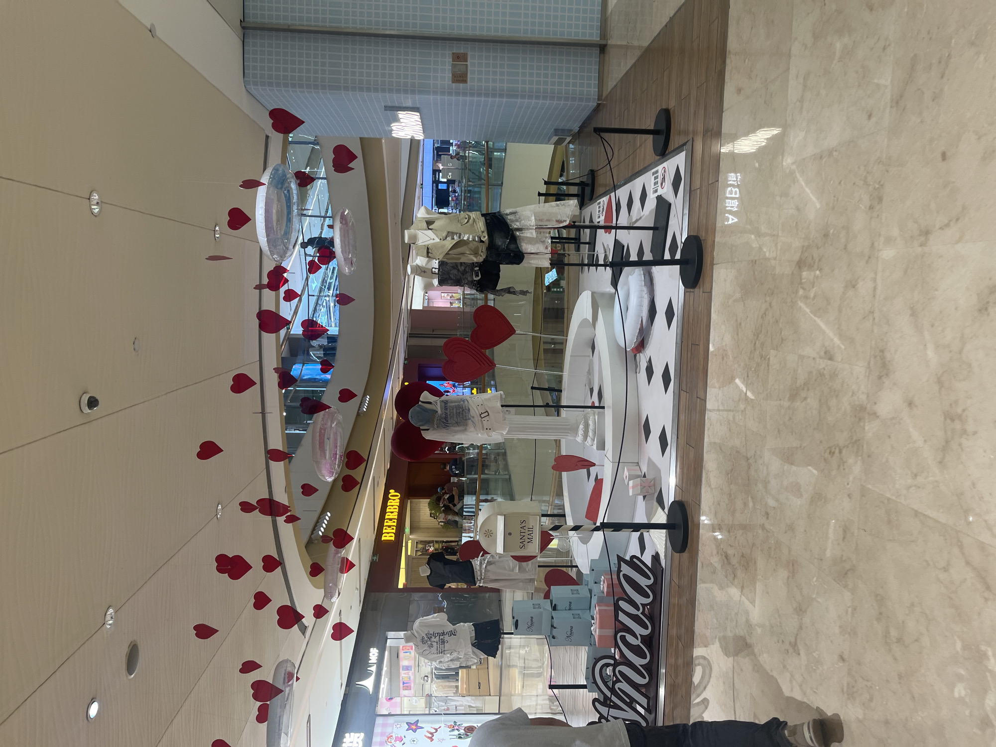 The image depicts an indoor display in a shopping mall, likely in the Yuzhong District, China, decorated with numerous red heart-shaped ornaments suspended from the ceiling. The display features several mannequins dressed in fashion apparel, arranged around a central circular platform. Surrounding the platform are gift boxes, a mailbox labeled "SANTA'S MAIL," and a large, stylized sign that appears to spell "Imnoya." The floor is a polished marble, reflecting the overhead lights and decorations. In the background, other shops and mall patrons are visible, suggesting a commercial environment. The overall scene conveys a festive or promotional atmosphere.