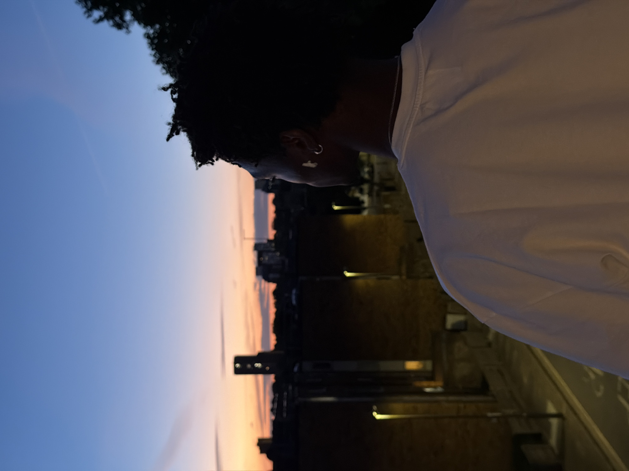 A person with dark, curly hair is viewed from behind, looking out at a city skyline during dusk. They are wearing a white t-shirt and a silver chain necklace. A silver hoop earring is visible in their left ear. The background shows buildings, including a tall tower with illuminated windows, silhouetted against a gradient sky of deep blue transitioning to orange and pink near the horizon. Streetlights are visible along a street below. The scene suggests an urban setting, likely in London, United Kingdom, given the architectural style and the characteristic skyline.