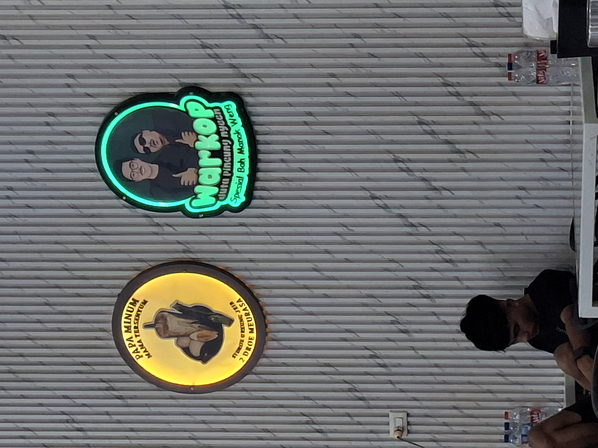 The image shows two illuminated signs on a vertical striped wall. The sign on the left is circular with a yellow glow and depicts a beverage and some food items. Text above reads "PAPA MINUM MAMA TERSEYUM" and below it, "STROKE 2 DROE MEURASA". The sign on the right is a rounded rectangle with a green glow and features a cartoon illustration of two men. Text within the green glow reads "warkop" and "duta pincung nyeeen spesial Bah Manok Weng".
In the foreground, a young man with dark hair and a black t-shirt is sitting with his arms crossed, looking down. He is seated at a white table. To the left of the man, there are three bottles of water on the floor. To the right of the table, there are two more bottles of water and a white tissue. The wall has a textured, ribbed pattern with dark grey and white marbling. The environment suggests an indoor setting, possibly a cafe or restaurant. The text on the signs indicates that the establishment is located in Sigli, Indonesia, a city in the Aceh province.
