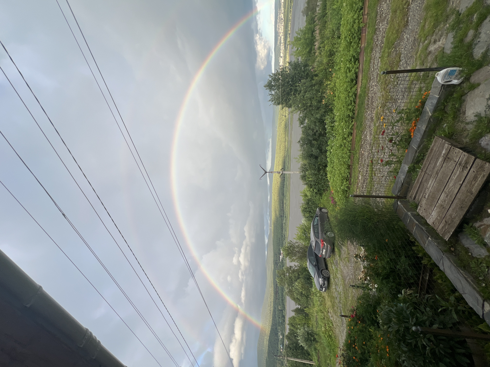 The image captures a wide-angle view of a landscape under a cloudy sky, dominated by a prominent double rainbow. In the foreground, a section of a roof overhang is visible at the top, with power lines running horizontally across the frame. Below the power lines, a vibrant rainbow arches across the sky, with a fainter secondary rainbow visible above it.
The middle ground features two parked cars, a dark sedan and a gray wagon, situated on a gravel area. Behind the cars, lush green vegetation, including trees and bushes, forms a dense backdrop. A body of water, likely a river, stretches across the scene, separating the near landscape from a distant, tree-covered mountain range. In the far distance, a town or city is faintly visible on the horizon.
The foreground also includes a small garden area with blooming flowers, a wire fence, and a rustic wooden structure. A white plastic container is present on the ground near the fence. The overall impression is of a natural scene, likely captured after a rain shower, with man-made elements such as the power lines and cars integrated into the environment.
This scene is consistent with the natural beauty found in and around Zlatoust, Russia, a city known for its picturesque landscapes, including the Taganay National Park. The presence of a rainbow is a common occurrence in regions with frequent rainfall and varied weather patterns.