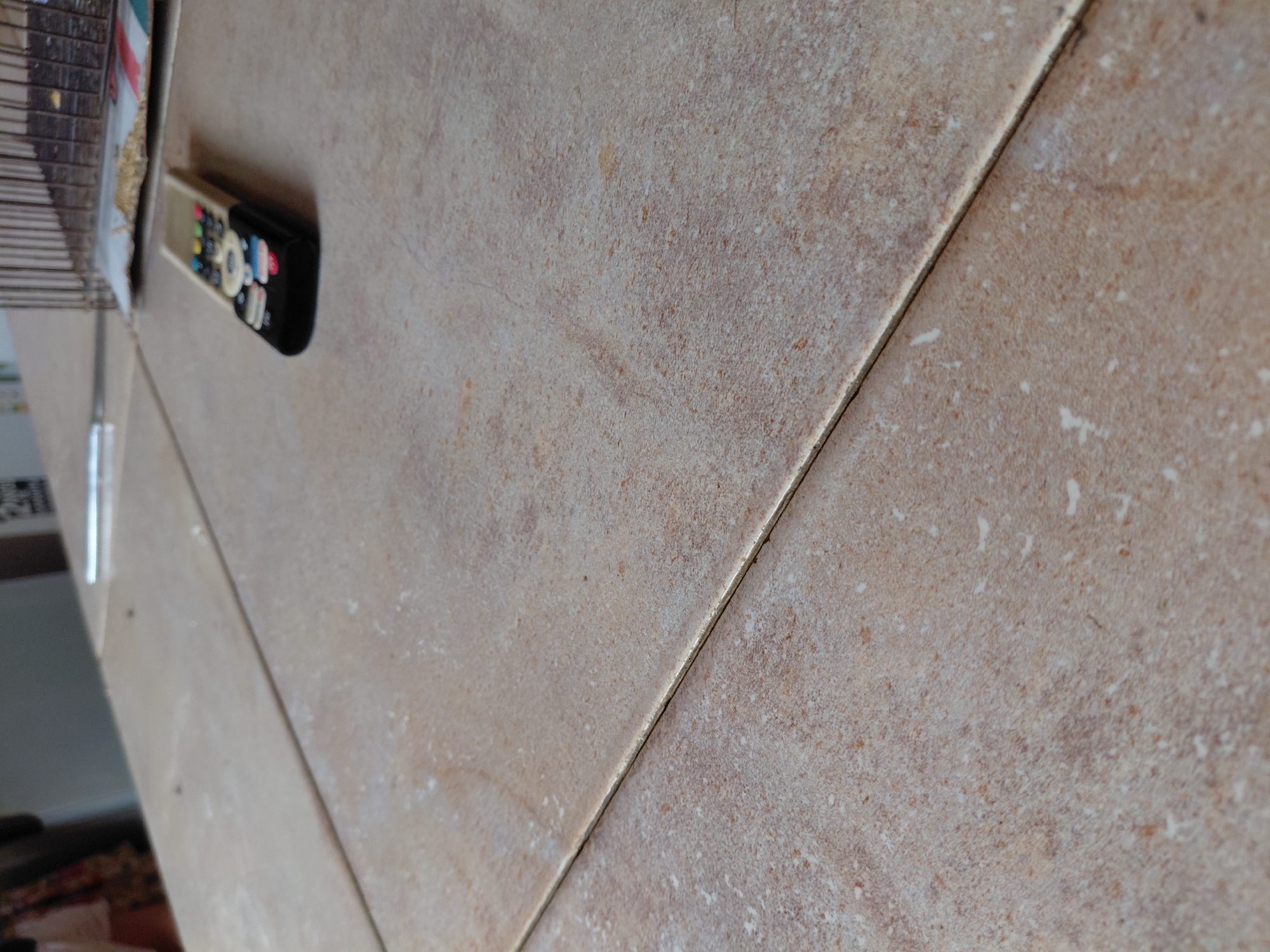 The image provides a close-up, high-angle perspective of a tiled surface in what appears to be an indoor setting in Santa Ana, Venezuela. The tiles are a light beige or sandy brown hue, characterized by subtle patterns and small, scattered specks of darker brown and white, giving them a natural stone-like appearance. A clear, slightly textured grout line runs diagonally across the frame, dividing two large tiles.
Resting on the upper-left tile is a dark-colored remote control. It features an assortment of colorful buttons, including red, blue, yellow, and green, alongside a prominent white circular control pad. Text or a brand logo is visible on the lower portion of the remote, but it is too blurry to be read distinctly. To the far left of the frame, partially obscured and out of focus, are faint hints of other objects, possibly a stack of items like papers or magazines, and a slender, reflective vertical element, potentially a table leg or part of a structure.
The scene is brightly lit, suggesting it is daytime, with natural light casting an even glow over the surface. No people are present, and no particular activity is taking place; the remote control simply rests on the tiles. The overall impression is that of a calm, everyday domestic or casual environment. No other discernible text is visible within the image.