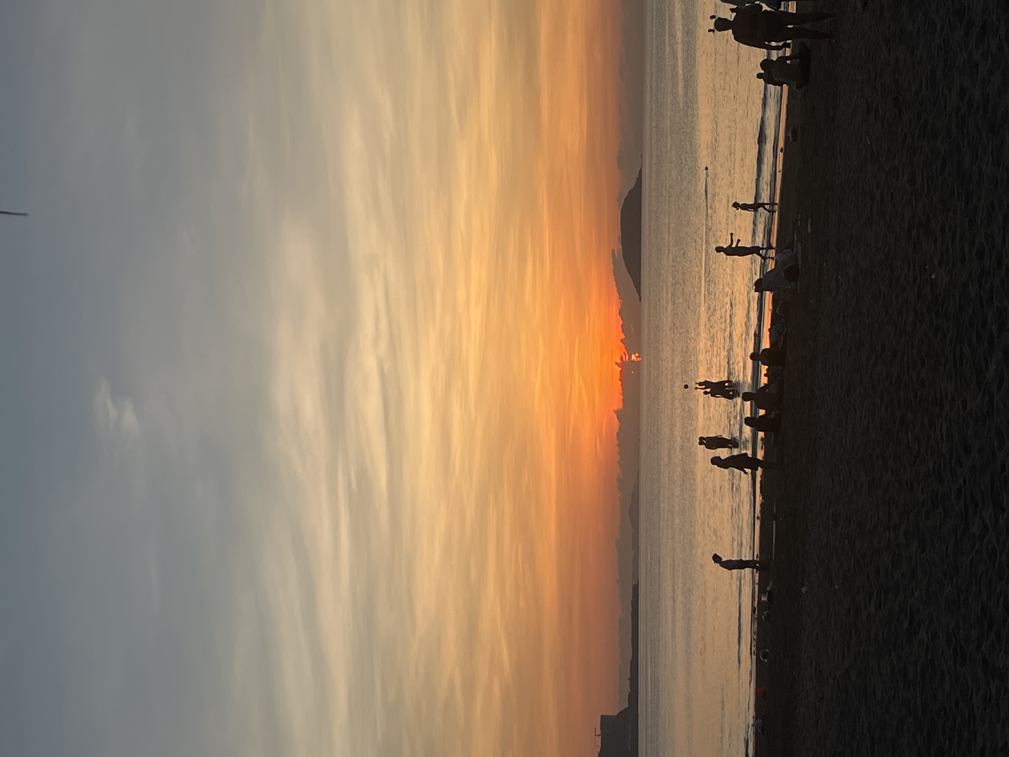This is an image of a beach scene at sunset in Bac Nha Trang Ward, Vietnam. The sky is a gradient of colors ranging from pale blue to orange and yellow near the horizon. The sun is setting, creating a bright glow. Silhouetted figures of people are visible on the beach, some standing and some interacting near the water's edge. A dark landmass or island can be seen in the distance on the water. The beach is composed of dark sand.