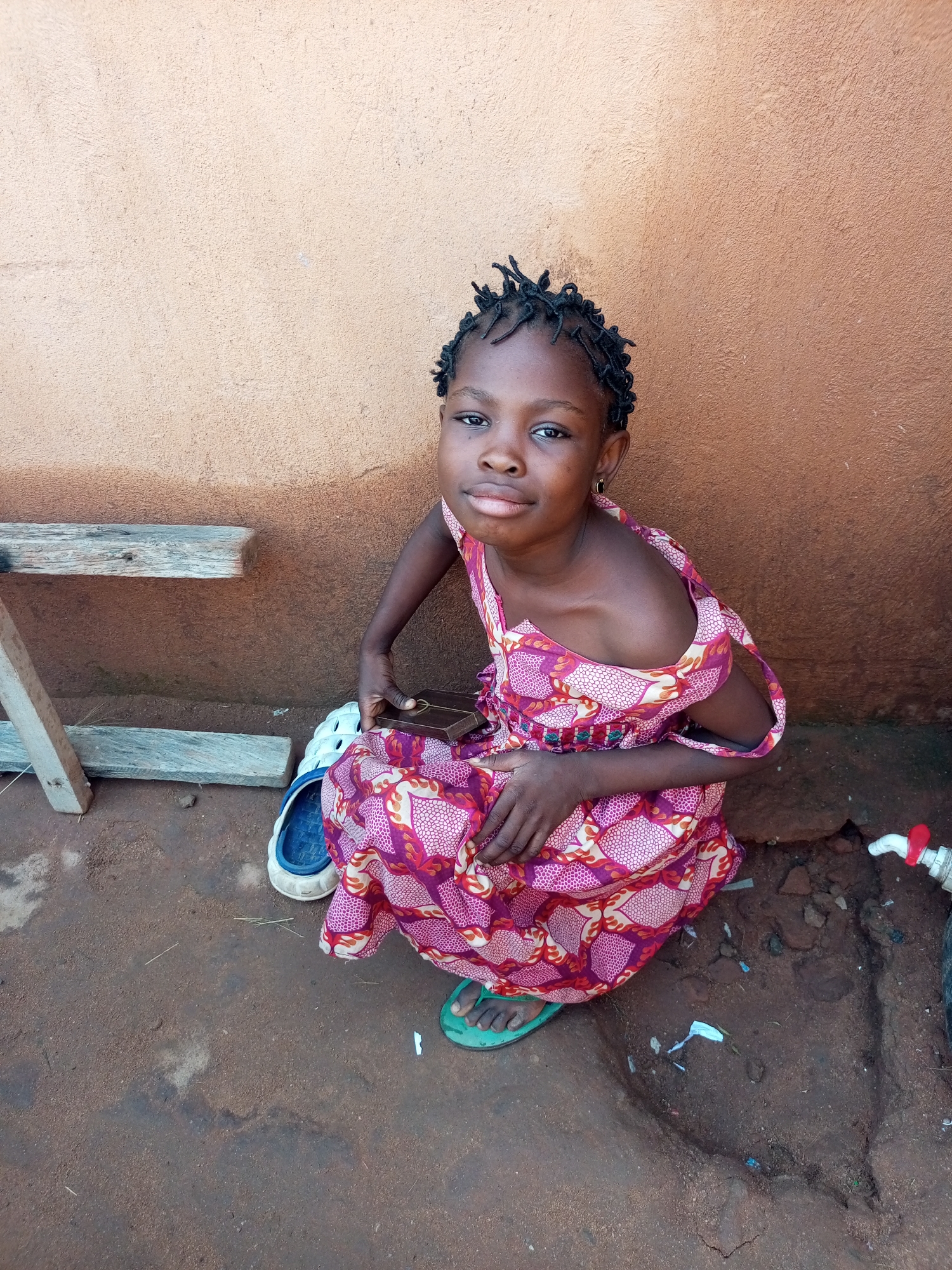 A young girl with dark skin and braided hair sits on the ground in front of a textured, ochre-colored wall. She wears a pink and red patterned dress with a light pink lining. Her right arm is bent, and she holds a small, dark rectangular object in her hand, possibly a book or a phone. She is wearing green flip-flops on her feet, and a pair of white and blue sandals are near her. To her left, there are two wooden planks supported by a vertical wooden post. The ground is dirt and appears somewhat uneven. In the lower right corner of the image, a white tap with a red handle is visible. The scene appears to be an outdoor setting, likely in a residential area in Gombe, Nigeria, characterized by the building material and the general environment.