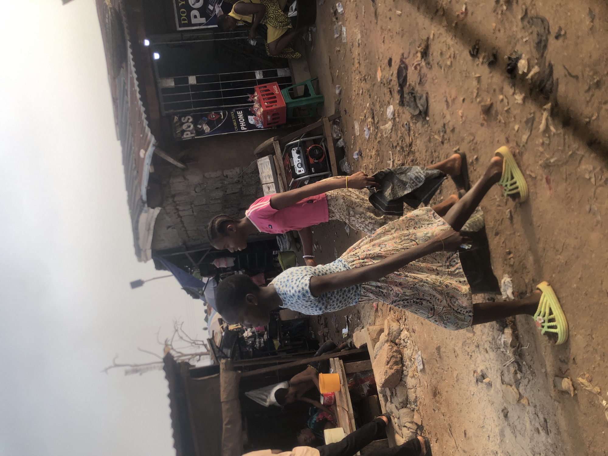 An outdoor scene in Onitsha, Nigeria, shows a bustling, unpaved market or street area under an overcast sky. The ground is dusty and littered with various debris, including plastic wrappers and small stones.
In the foreground, two young girls are walking or standing. One girl wears a pink t-shirt and a patterned skirt, carrying a dark-colored bag. The other girl wears a light blue and white patterned dress and distinctive bright lime green sandals. Their expressions are neutral as they move through the environment.
The background features several makeshift structures, likely market stalls or small shops, with corrugated metal roofs. A generator sits on a wooden stand near one of these structures. Nearby, a red Coca-Cola crate filled with items is visible, along with a green plastic stool. Other individuals are present in the background, some seated or standing, contributing to the sense of a busy daily scene.
Visible text on signs includes "DO YOU POS" and "CHARGE YOUR PHONE," indicating services offered at one of the stalls. Another sign simply says "POS." The overall impression is one of everyday life in an informal urban or semi-urban commercial area.