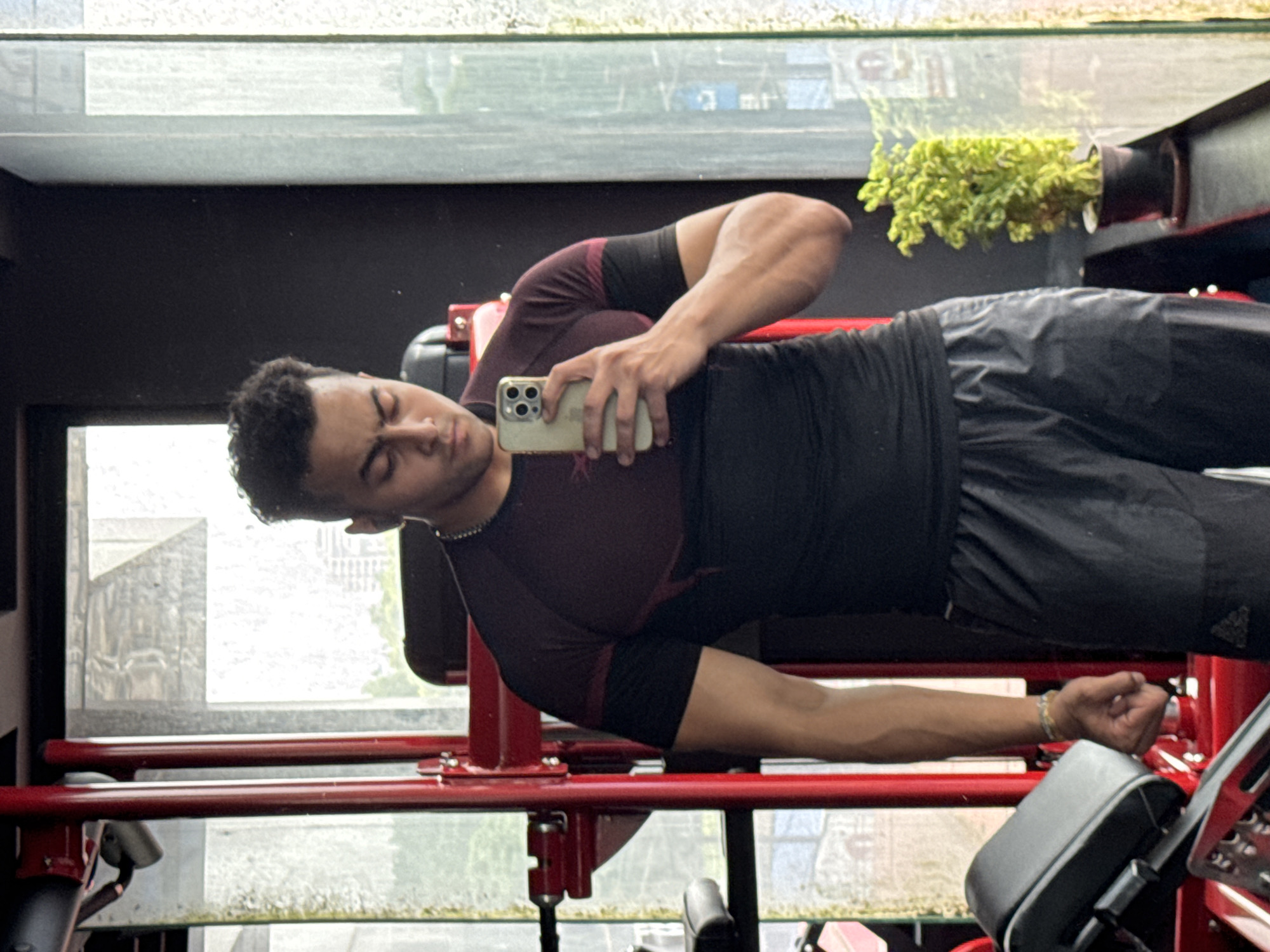 A young man with dark, curly hair takes a selfie in a gym in Kolkata, India. He is wearing a dark t-shirt with burgundy accents and dark athletic shorts. He holds a smartphone in his right hand, angled to capture his reflection. The gym equipment, predominantly red, is visible behind him. A large window to his right offers a view of the city, with buildings and greenery apparent. A potted plant sits on the windowsill. The lighting suggests it is daytime, possibly overcast.