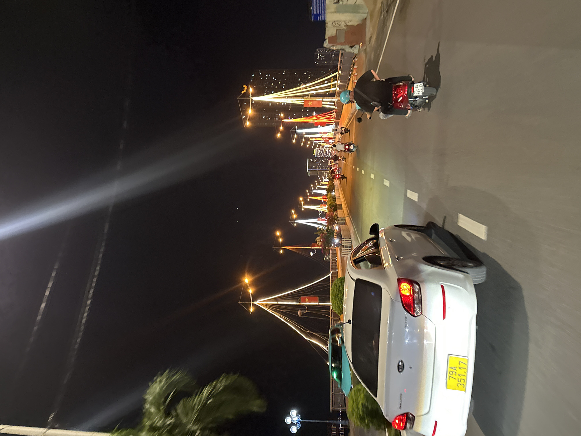 This is a night-time shot of a bridge in Nha Trang, Vietnam. The bridge is adorned with numerous lights, creating a festive atmosphere. Several vehicles, including cars and motorcycles, are visible on the road. In the foreground, a white car is seen from behind, its license plate reading "79A 351.17". Further ahead, a red motorcycle with two riders is traveling in the same direction. Other motorcycles and a turquoise car are also present on the bridge. The background features illuminated buildings and palm trees, indicating an urban coastal setting. The overall impression is that of a lively city night scene.