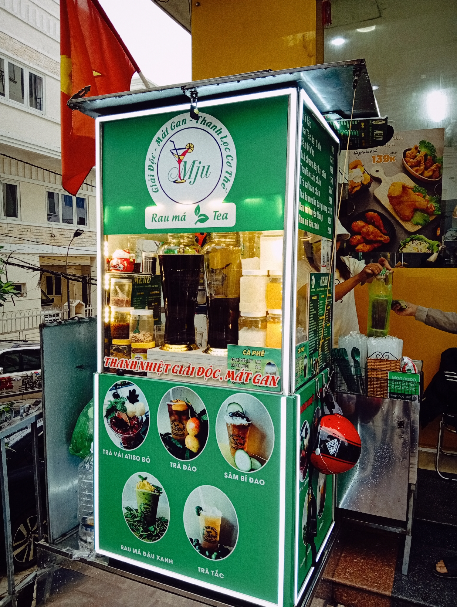 A brightly lit, green-themed street vendor stall, bearing the brand "Mju," stands on a sidewalk in what appears to be Da Lat, Vietnam. The stall specializes in teas, coffees, and detoxifying drinks, prominently featuring "Rau má Tea" (Centella Asiatica Tea). The top sign reads "Giải Độc - Mát Gan - Thanh Lọc Cơ Thể" (Detox - Cool Liver - Body Cleansing).
Inside the stall, large clear dispensers hold various dark and light liquids, likely pre-made drinks, alongside jars of ingredients. The stall's front and right side feature illustrated menus with drink options such as "TRÀ VẢI ATISO ĐỎ" (Red Artichoke Lychee Tea), "TRÀ ĐÀO" (Peach Tea), "SÂM BÍ ĐAO" (Winter Melon Tea), "RAU MÁ ĐẬU XANH" (Centella Asiatica with Green Bean), and "TRÀ TẮC" (Kumquat Tea), with prices ranging from 10K to 20K VND. Other offerings include "CÀ PHÊ SỮA DỪA" (Coconut milk coffee) and "CÀ PHÊ MUỐI" (Salt coffee). A sign on the lower front reiterates "THANH NHIỆT GIẢI ĐỘC, MÁT GAN."
On the right side of the stall, a vendor, partially visible in a white shirt and cap, is serving a green beverage in a plastic bottle to a customer whose hand is extended, suggesting a transaction. Nearby, stacks of plastic cups, ice, and green utensils are neatly arranged. A red helmet hangs from the side of the kiosk. Behind the vendor, a large poster advertises fried food items for "139K," and a small sign indicates "Món ngon đã có trên GrabFood" (Delicious dishes available on GrabFood).
In the upper left corner, a red Vietnamese flag with a yellow star is visible. The background reveals white, multi-story buildings and parked cars, typical of an urban setting. The overall scene captures a lively street vending operation during daytime.