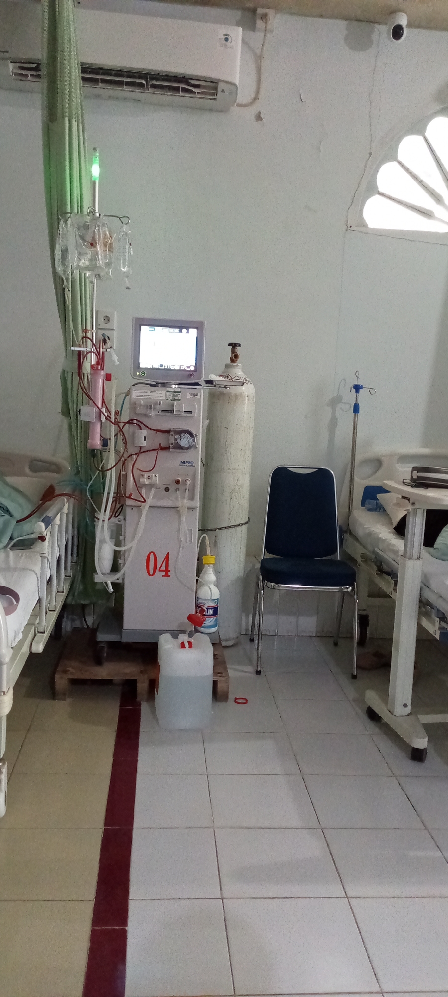 This media file captures a medical treatment room, likely a hemodialysis unit, in Lhokseumawe, Indonesia. The central focus is a white NIPRO hemodialysis machine, labeled "04" in red, with a monitor displaying data positioned on top. Various red and white tubes, filters, and medical components are connected to the machine. An IV stand with several fluid bags hangs to the left of the machine, emitting a green light from its top.
To the right of the dialysis machine, a tall, light-colored oxygen tank stands secured by a chain. Below the machine, a white plastic container and a larger clear plastic jerry can rest on a wooden pallet. A white bottle, identifiable by the "SOKLIN" brand label, is placed next to the machine.
Two hospital beds are visible; one is partially obscured on the left, while another on the right has a movable bedside table. A dark blue upholstered chair with a metal frame is positioned between the oxygen tank and the right hospital bed. Above the IV stand, an air conditioner is mounted on the white wall.
The room features white tiled flooring, bisected by a distinctive dark red tile strip. The walls are painted white but show signs of wear, including cracks and discolored patches. An arched, multi-pane window with white frames is visible in the upper right corner, and a small, white security camera is mounted high on the wall nearby.
The scene depicts a functional, clinical environment, equipped for medical procedures, specifically dialysis. No people are present, indicating the room is currently unoccupied or awaiting patients. The visible text includes "NIPRO" and "04" on the machine, and "SOKLIN" on the cleaning bottle.