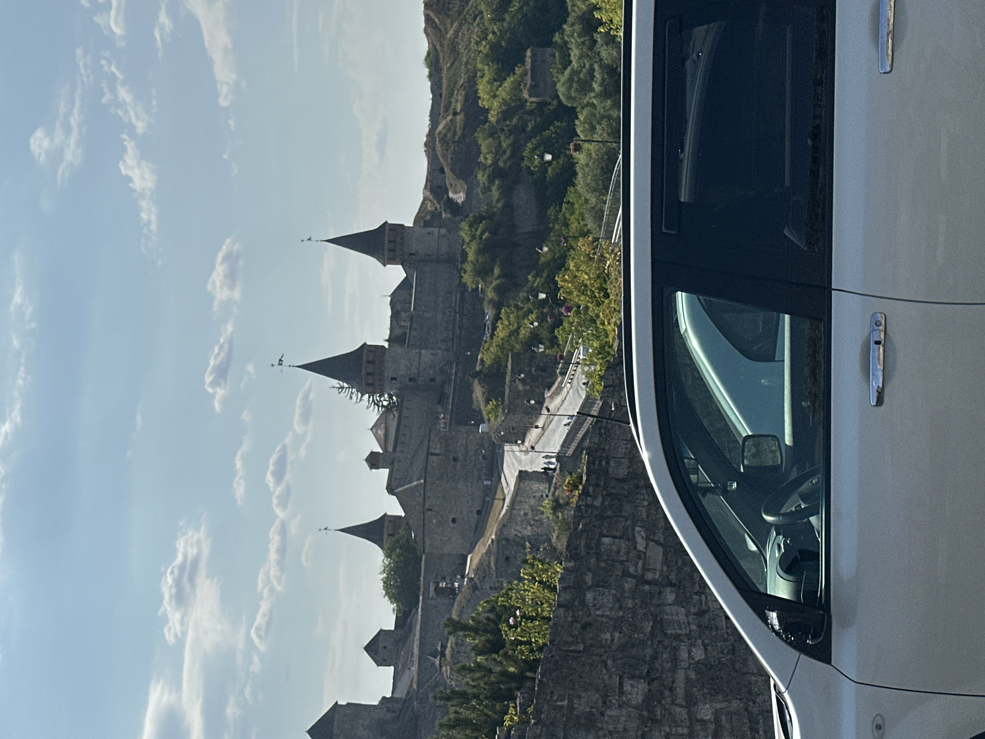 The image depicts a view of the Kamianets-Podilskyi Castle in Ukraine, taken from a vehicle. In the foreground, the white exterior of a car is visible, with its side door, window, and door handle in focus. The car's interior, including a steering wheel and dashboard, is partially visible through the driver's side window. In the midground, a stone wall and lush green vegetation line a pathway that leads towards the castle complex. The castle, an impressive medieval fortress, stands prominently on a hill, characterized by its stone towers and crenellated walls. Several figures, appearing to be tourists, can be seen walking along the path and within the castle grounds. The background consists of a hazy sky with scattered clouds and more rolling green hills surrounding the historical site.