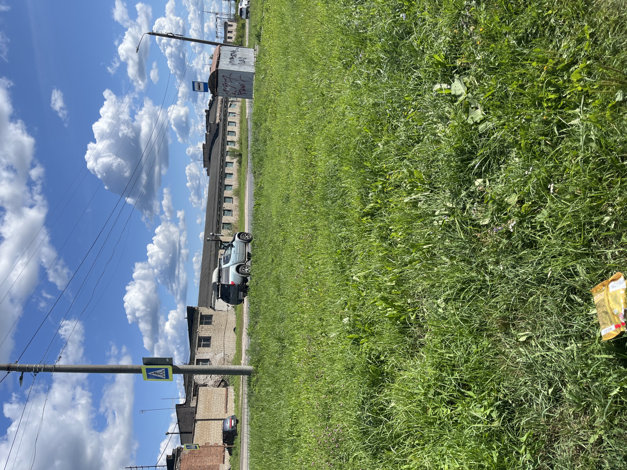 In a sunlit scene with a bright blue sky filled with puffy white clouds, a grassy area occupies the foreground. A yellow and black pedestrian crossing sign is mounted on a utility pole on the left side of the image, near a brick building. Further down the road, a light blue pickup truck with a roof rack drives past a long, low-rise brick building. To the right, a bus stop shelter with graffiti on its corrugated metal exterior stands beside the road, with another building visible in the background. The grassy area in the foreground is slightly overgrown and contains some small wildflowers. A crumpled yellow wrapper lies on the grass in the lower left corner of the frame.