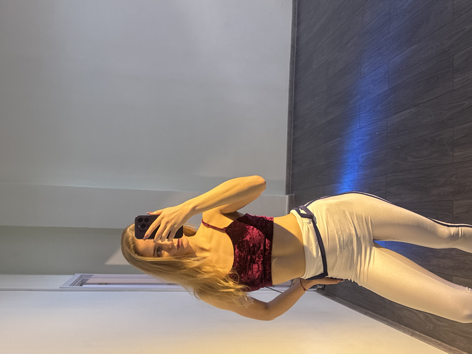 A full-body mirror selfie depicts a young adult female in an Unknown location in Unknown, Unknown. The subject, possessing long blonde hair, is partially obscured by a dark-colored smartphone held in her right hand, which covers her upper face. Her left hand rests on her hip. She is wearing a dark red velvet crop top with thin straps and white athletic leggings with dark blue vertical stripes and a patterned waistband. The indoor environment consists of light-colored walls and dark wooden flooring with baseboards. A door frame is visible on the left side of the reflection. The scene is illuminated by a warm, yellowish light source from the left, with a distinct linear blue light reflection visible on the floor to the right.
