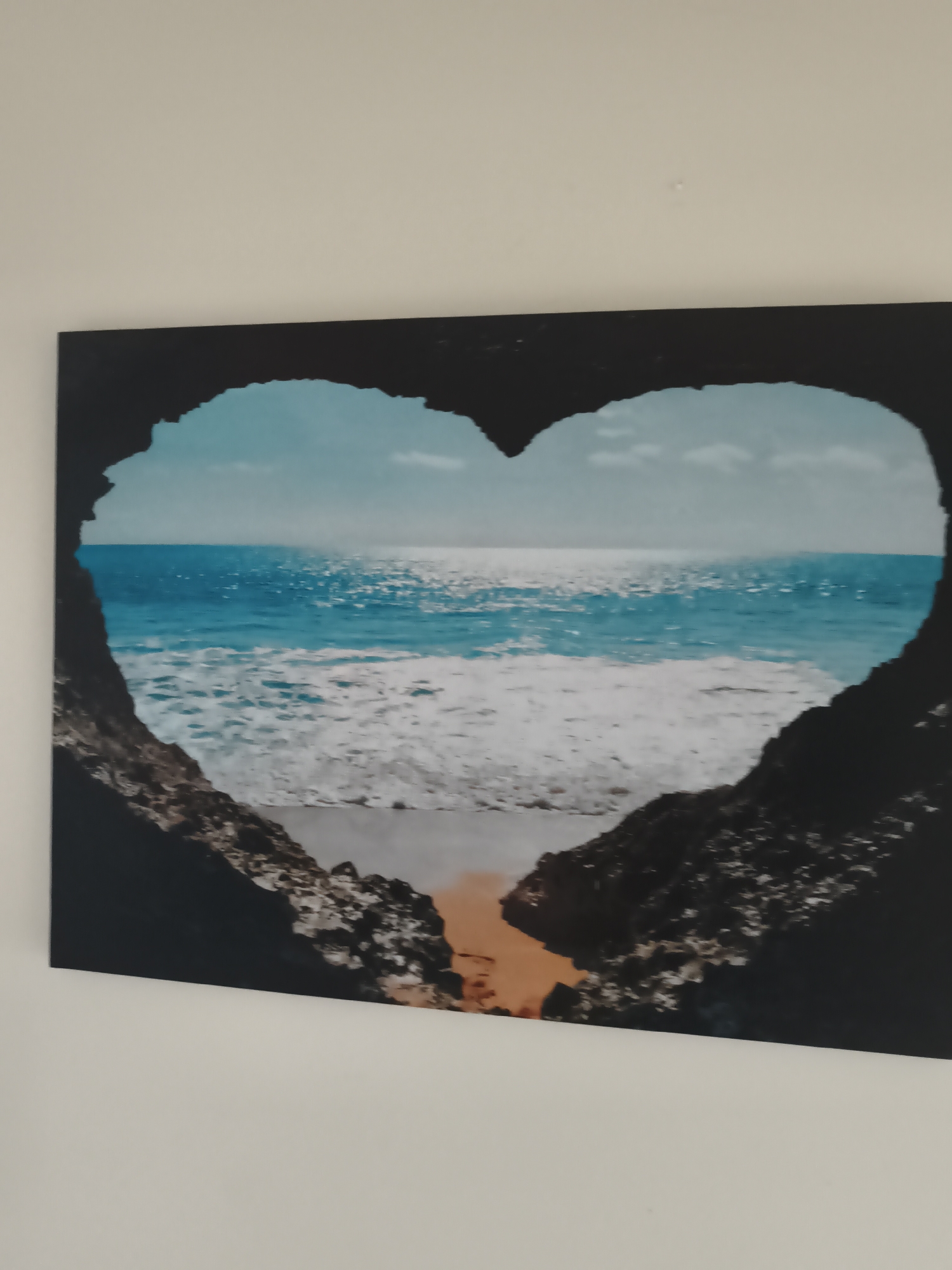 An image displays a piece of wall art, likely a canvas print or framed picture, mounted on a plain, light-colored wall. The artwork itself depicts a vibrant ocean scene framed by a natural, heart-shaped rock formation, suggesting a view from inside a cave or through a natural archway.
The heart-shaped opening is dark and rocky, creating a stark contrast with the brightly lit seascape beyond. The ocean is rendered in various shades of blue, with lighter aqua tones closer to the shore and a deeper blue extending to the horizon. Sunlight glints brightly on the water's surface, indicating a sunny daytime scene with clear weather. Waves are visibly crashing near the foreground, creating white foam. A small strip of sandy or light-colored beach is visible at the very bottom of the heart-shaped frame, just before the water. The sky above the ocean is a pale blue with a few subtle clouds.
The overall impression is one of tranquility and natural beauty, with the heart shape adding a romantic or symbolic touch. There is no specific activity or event taking place, and no people or animals are visible in the scene. No text is visible on the artwork or the surrounding wall. The wall itself is a uniform light beige or off-white, suggesting an indoor setting.