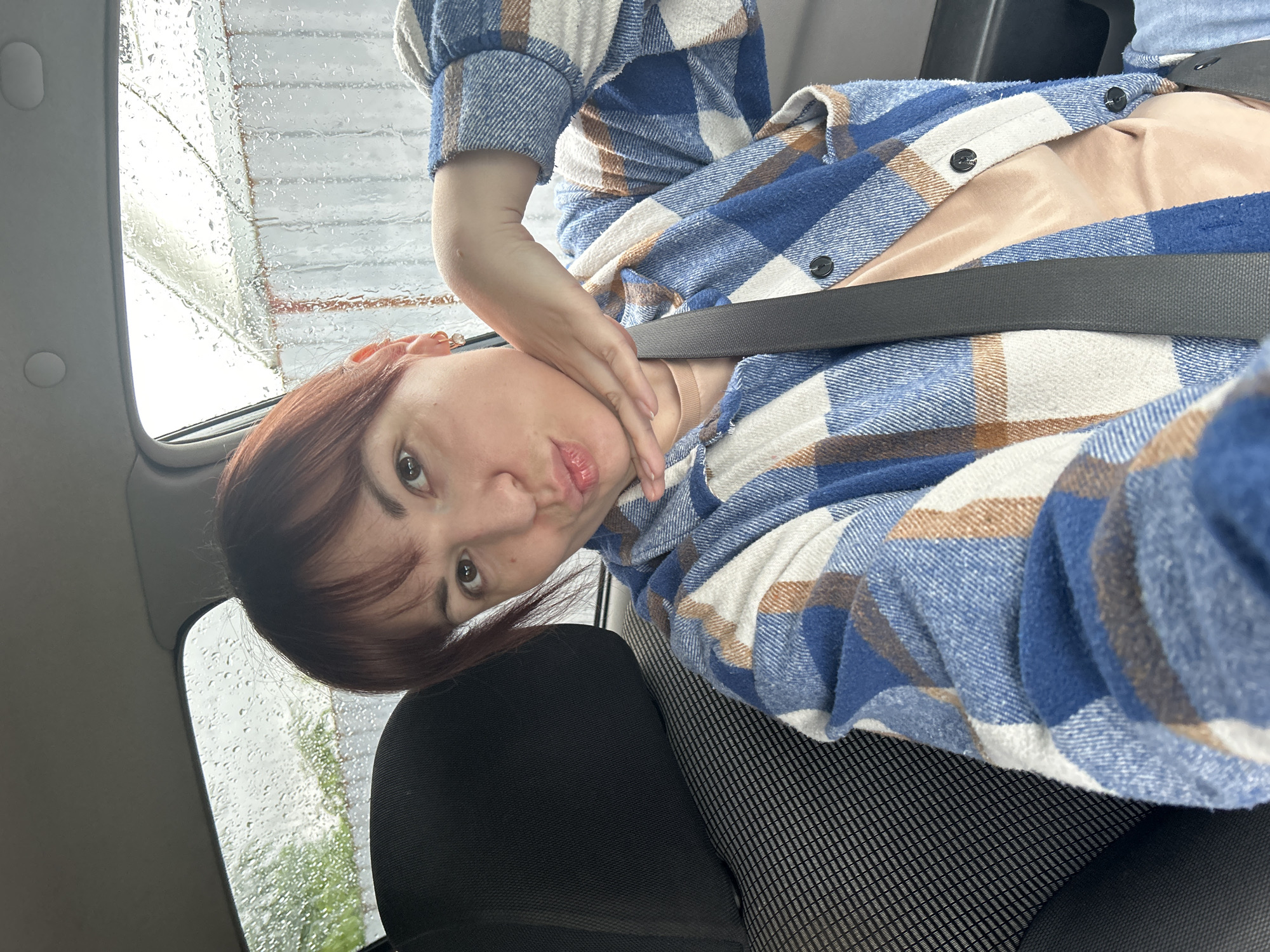 A woman with auburn hair wearing a blue plaid shirt with a peach-colored shirt underneath is sitting in a car with a seatbelt on. The car window has raindrops on it, blurring the outside view. The location is Фоки, Russia.
