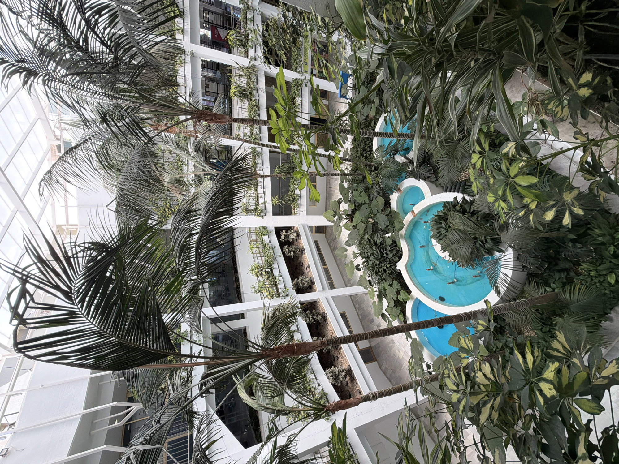 The image shows an indoor tropical environment in Arroyo de la Miel-Benalmádena Costa, Spain. The scene includes a multi-tiered turquoise fountain surrounded by dense foliage, including palm trees and various other plants. The architectural structure consists of a white building with tiered window boxes containing vegetation. The perspective is angled upward, capturing the building's height and the overhead light. No people are visible. The setting is an indoor atrium or courtyard.