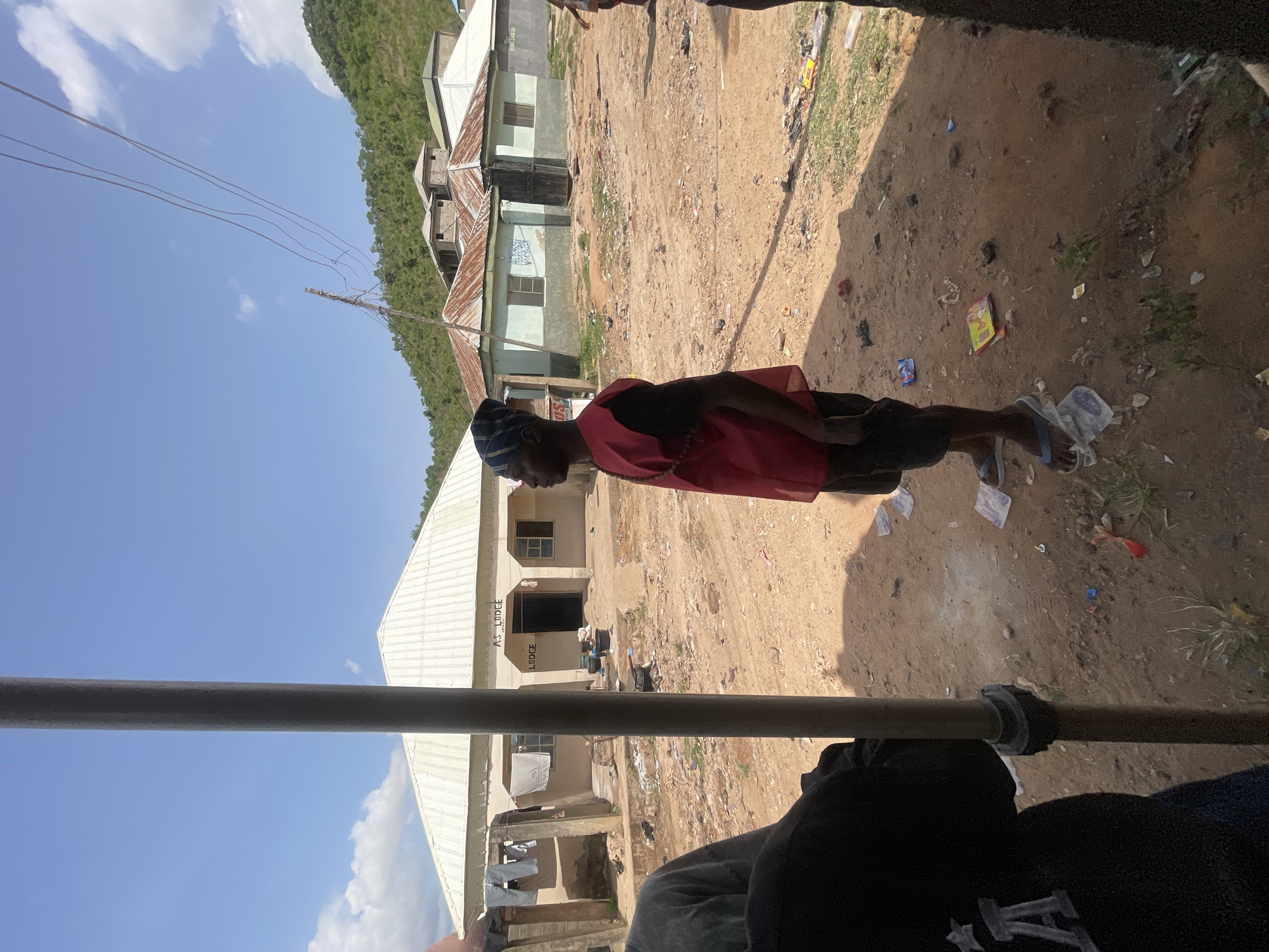 A medium shot, looking out from inside a vehicle, shows a man in a red garment standing on a dirt road outside AS Lodge in Lokoja, Nigeria. The man wears a blue patterned head covering, a red sleeveless vest or garment, shorts, and flip-flops. He appears to be looking at something to his left. The surrounding environment is littered with trash. A building identified as "AS Lodge" is visible with clothes hanging outside. Other buildings are visible in the background, along with a tree-covered hillside. Overhead power lines stretch across the sky. The sky is blue with some clouds. The foreground features a metal bar and part of the vehicle interior with a partially visible person.