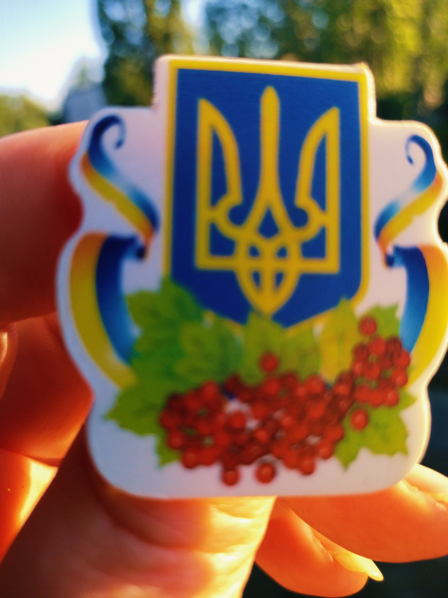 A close-up, blurred image shows a hand holding a sticker or magnet with a Ukrainian national symbol, the Tryzub (trident), on a blue background with a yellow border, and depicted with red berries and green leaves. The background is out of focus, showing green foliage and blue sky. The scene is illuminated by bright sunlight, casting shadows and highlights on the hand. The image is taken outdoors, likely in a park or garden area in Horishni Plavni, Ukraine, given the subject matter.