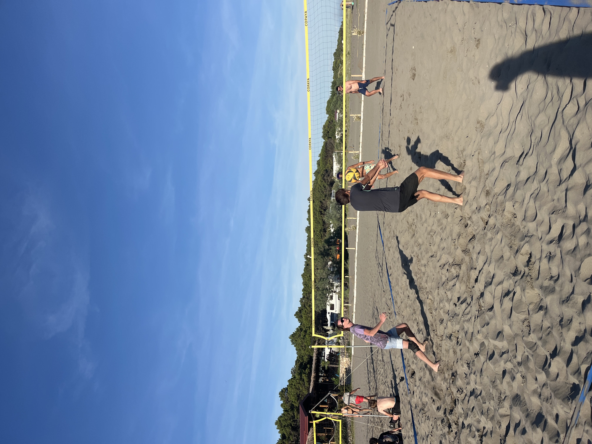 This image captures a beach volleyball game on a sandy court, likely in Pistula, Montenegro. The sky above is a clear, bright blue with a few wispy clouds. A yellow volleyball net stretches across the court, with the word "COPAYA" visible on it. On the court, four individuals are actively engaged in the game.
In the foreground, a person wearing a purple t-shirt and light-colored shorts is in motion, facing away from the camera and towards the net. To their left and slightly behind, a person in a grey t-shirt and dark shorts is in the process of hitting a yellow volleyball. Further behind them, another person in patterned shorts is observed. On the opposite side of the net, a person in dark blue shorts is standing, looking towards the game.
The beach itself is covered in sand, with visible footprints and rippled patterns from the wind. In the background, there is a line of green trees, a building with a red roof and thatched elements, and several parked camper vans. The overall scene depicts a lively recreational activity on a sunny day at the beach.