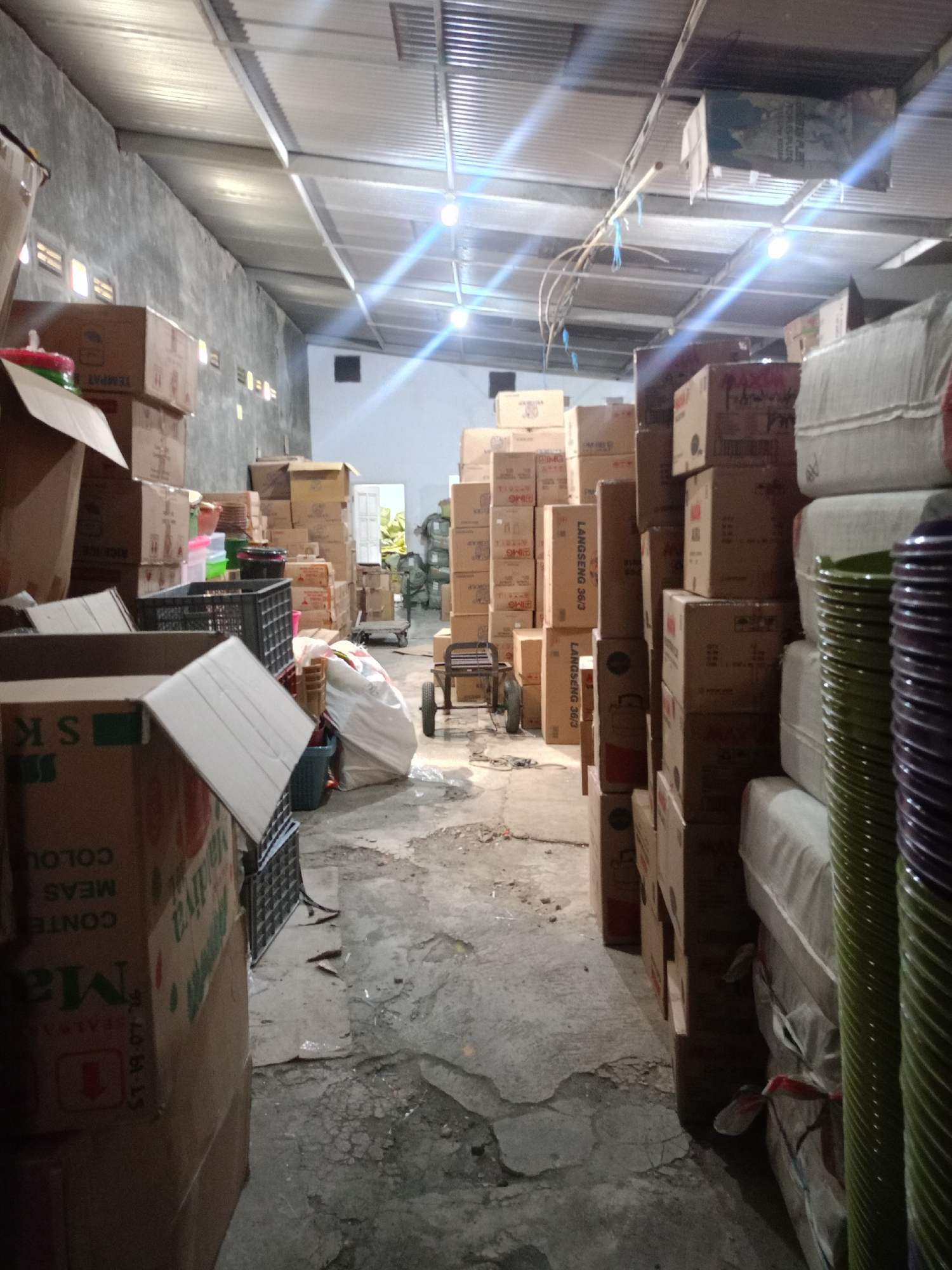 The image shows the interior of a warehouse located in Batu, Indonesia. The warehouse is filled with stacks of cardboard boxes, some of which are labeled with text. There are also several plastic crates, a large white plastic bag, and a stack of what appear to be colorful plastic plates or bowls on the right side of the image. A small hand cart with two wheels is positioned in the center of the aisle. The ceiling is made of corrugated metal, and fluorescent lights are visible. The floor is concrete and shows signs of wear and tear. The overall impression is that of a storage facility.