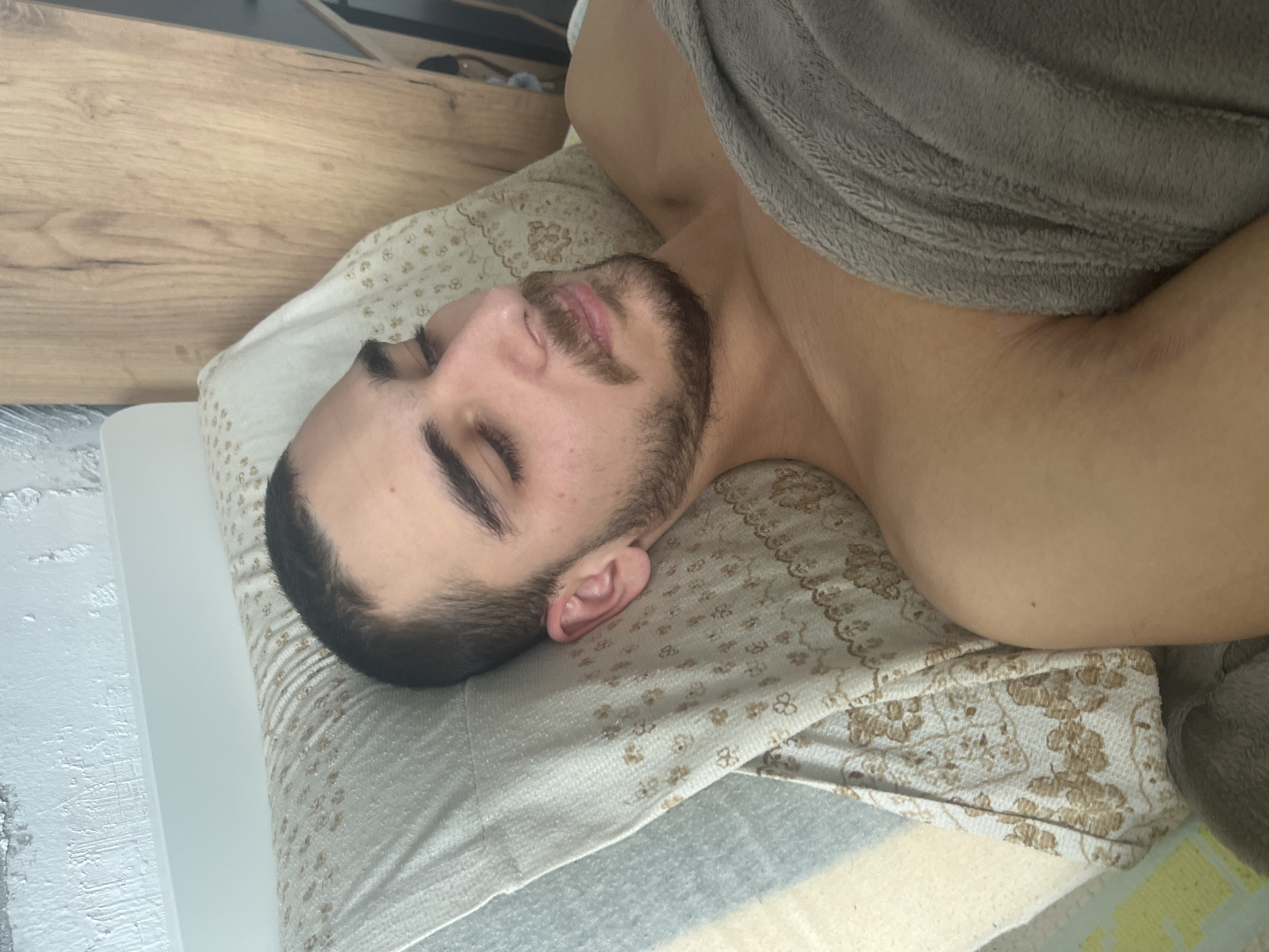 A man is lying on his side on a pillow, covered with a grey blanket. He has dark hair, a beard and mustache, and his eyes are closed. The pillow has a white and gold patterned cover. The setting appears to be indoors, possibly in a bedroom. It is not possible to determine the exact location in Varna, Bulgaria, or other specific details from the image provided.