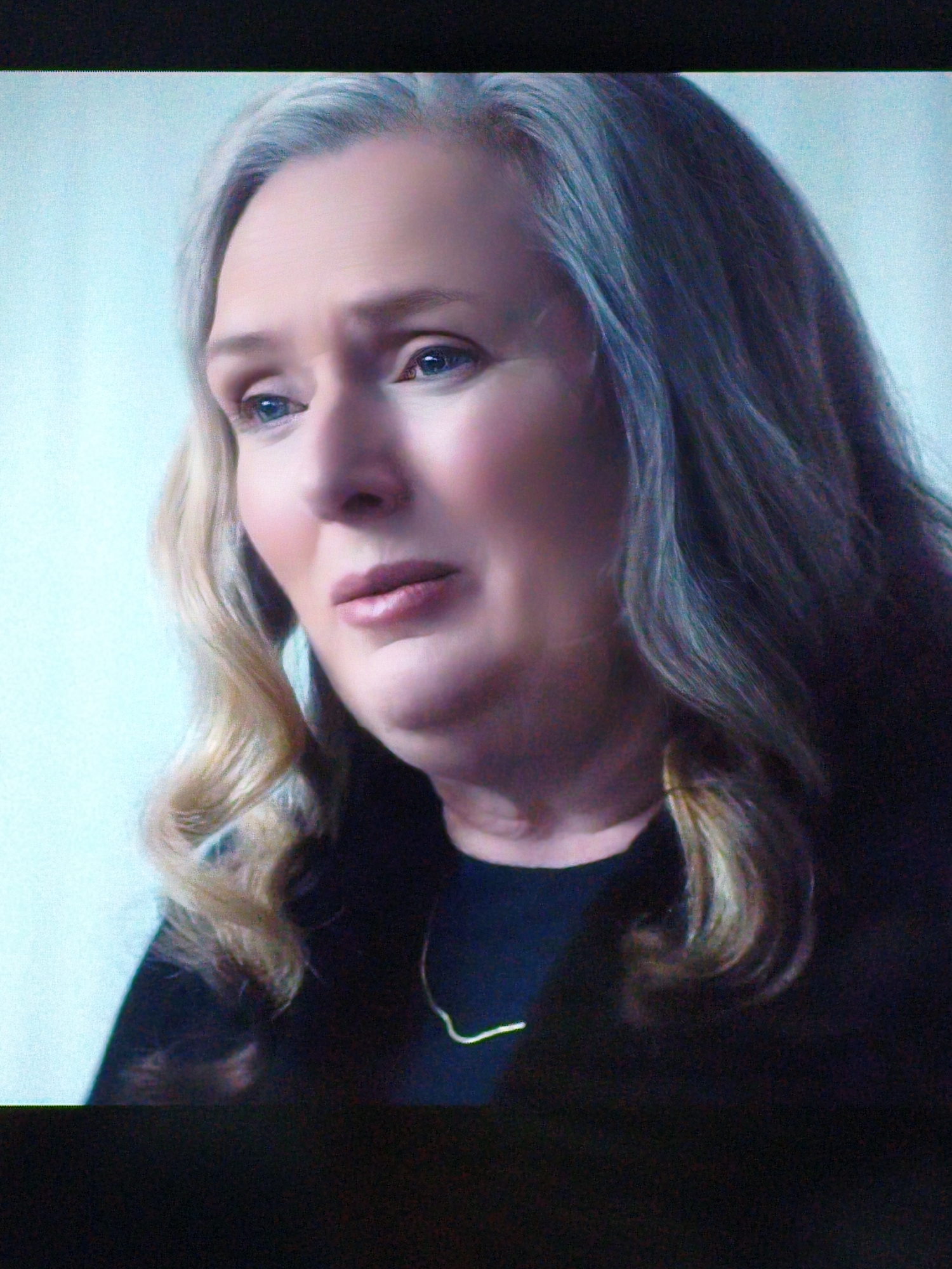 The image shows a medium close-up of an older woman with fair skin, blue eyes, and blonde hair with grey streaks. She is wearing a black top and a thin, gold necklace. Her face is turned slightly to the left, and she has a sad or concerned expression. The background is a plain, light blue. There are no identifiable landmarks or elements that would indicate a specific location in Vladivostok, Russia. The image is a still from a video, as evidenced by the black bar at the top and bottom which appears to be part of a screen recording.