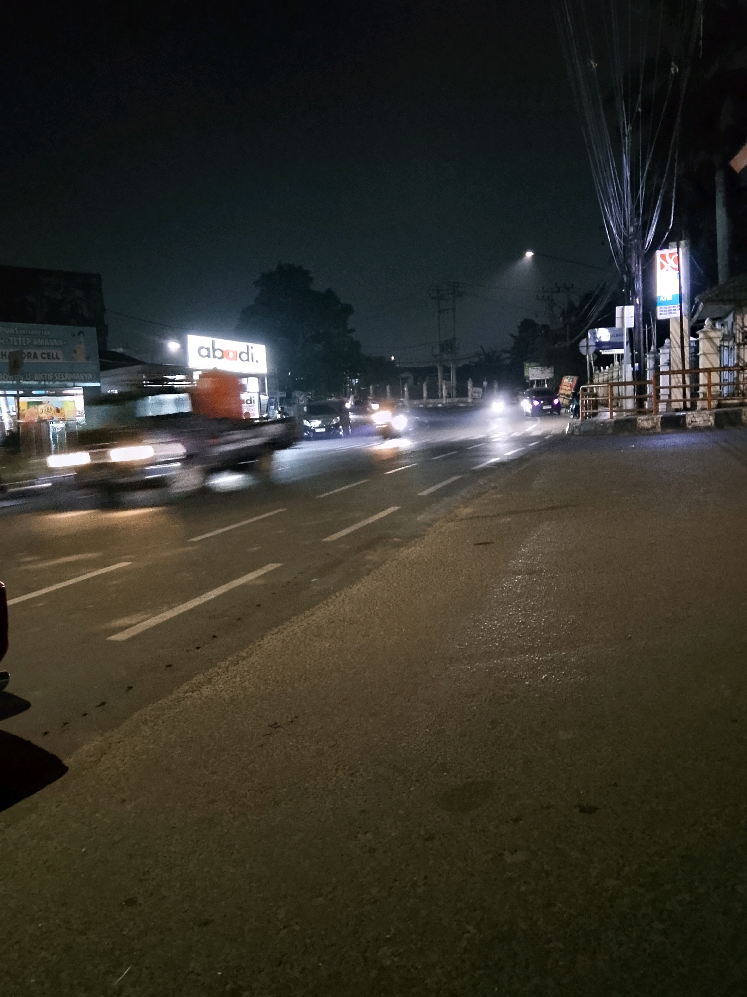 The image depicts a street scene at night in Palembang, Indonesia, likely on a humid evening given the reflections on the asphalt. In the foreground, the road surface is dark and appears wet, with white lane markings visible. To the left, a portion of a dark-colored vehicle's shadow is cast on the road.
In the middle ground, a blurred light-colored truck with an orange container on its bed is moving from left to right, its headlights creating streaks of light. Adjacent to the truck, a car with its headlights on is also moving. Further down the road, additional blurred vehicles and their lights indicate traffic flow.
On the right side of the road, a brightly lit sign with the word "abadi." in red and white lettering is prominently displayed above a storefront. Next to the "abadi." sign, a pole with numerous electrical wires is visible, extending upwards into the dark sky. A white fence with reddish-brown horizontal railings runs along the side of the road. Beyond the fence, structures and trees are dimly visible against the night sky. The overall atmosphere is that of a busy urban street at night, with the illumination from signs and vehicle lights cutting through the darkness.