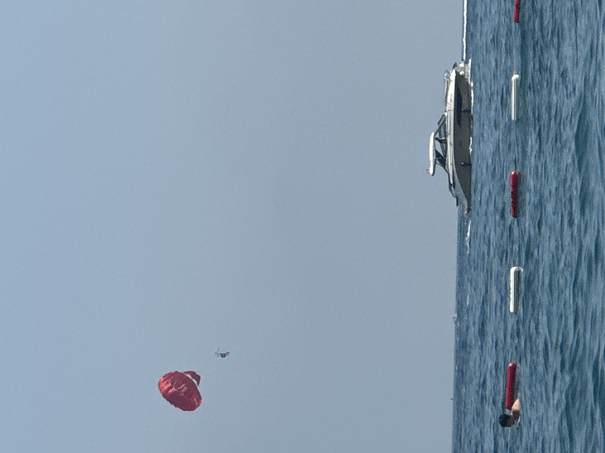 The image shows a bright, clear day with a calm blue sea. In the sky, a red parasail with a person suspended beneath it is visible, along with a distant boat. Closer to the water, a motorboat with several occupants is moving across the sea. In the foreground, a person is swimming in the water, accompanied by a red float and a white marker buoy. The scene suggests a recreational setting in a coastal area. The city of Boreti, Montenegro, is a coastal town known for its beaches and water activities, which aligns with the visual elements in the image.