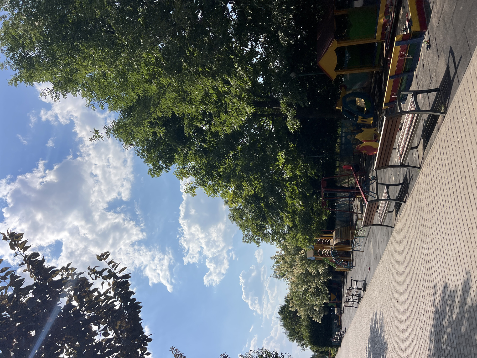 The image depicts a paved walkway in a park-like setting, with a playground visible to the right. The walkway is made of light-colored bricks or pavers, arranged in a uniform pattern. Several empty wooden benches with metal frames are placed along the edge of the walkway, facing the playground.
The playground area features a variety of equipment, including slides, swings, and a sandbox. The structures are colorful, with shades of yellow, red, and green. The ground of the playground appears to be covered with a dark rubberized surface.
Tall, leafy green trees line the background, creating a dense canopy that partially obscures the sky. The sky itself is blue with scattered white, fluffy clouds. Sunlight streams through the leaves of a dark-leaved tree in the upper left portion of the image, casting some light rays.
The scene appears to be empty of people, suggesting a quiet moment in the park. The overall impression is one of a well-maintained outdoor public space on a sunny day. The location is identified as Fontanka, Ukraine.