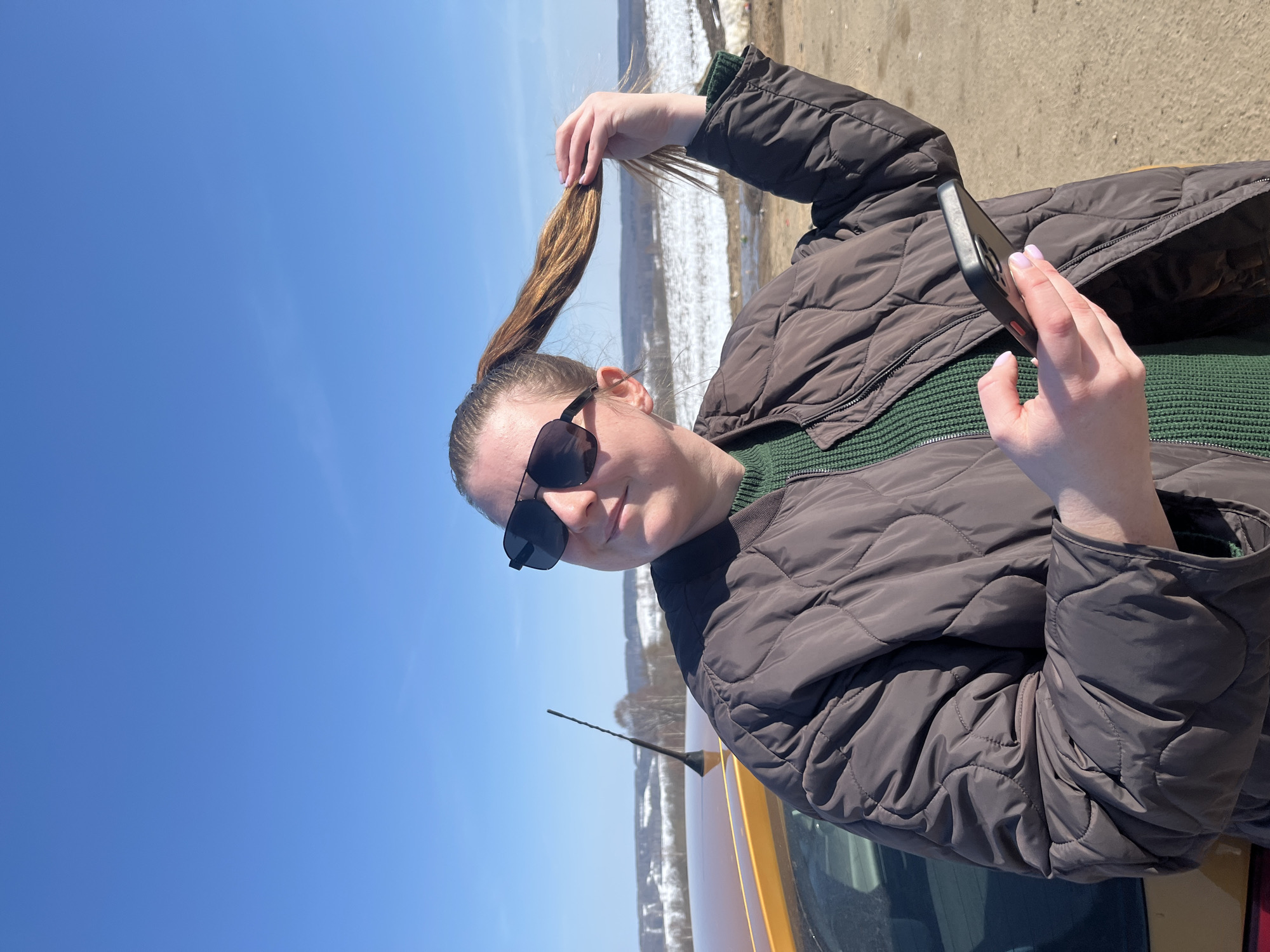 An adult female subject is depicted outdoors in Zubovo, Russia. She stands angled to the viewer's left, partially obscuring the rear section of a yellow vehicle. She wears dark, rectangular sunglasses, a dark green ribbed sweater, and a dark brown quilted bomber jacket. Her light brown hair is styled in a high ponytail, which she holds with her right hand. In her left hand, she holds a black-cased smartphone. The subject's head is turned towards her left, and she displays a slight smile. The background features a wide, undulating landscape with patches of white snow and visible bare trees, extending to a distant horizon under a clear, bright blue sky. The immediate ground surface appears to be bare earth or dirt.