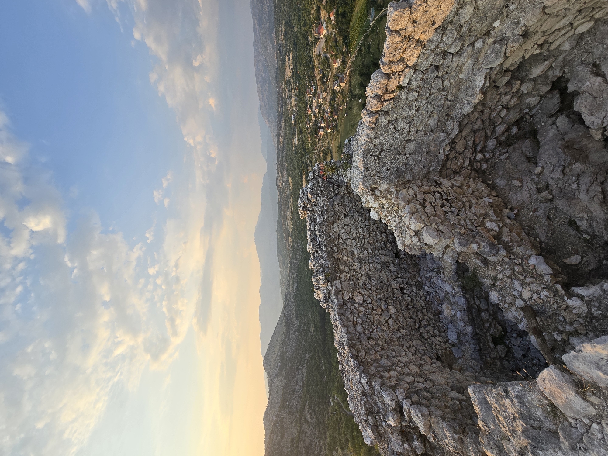 The image depicts a view from the ruins of Medun fortress in Medun, Montenegro. In the foreground, weathered stone walls of the ancient fortress are visible, showing evidence of decay and time. Beyond the ruins, a valley unfolds, dotted with houses and greenery, suggesting a rural settlement. The distant landscape is characterized by rolling hills and mountains, partially obscured by a hazy atmosphere. The sky above is a mix of blue and soft, warm-toned clouds, indicative of either sunrise or sunset. The overall scene conveys a sense of history, tranquility, and the vastness of nature.