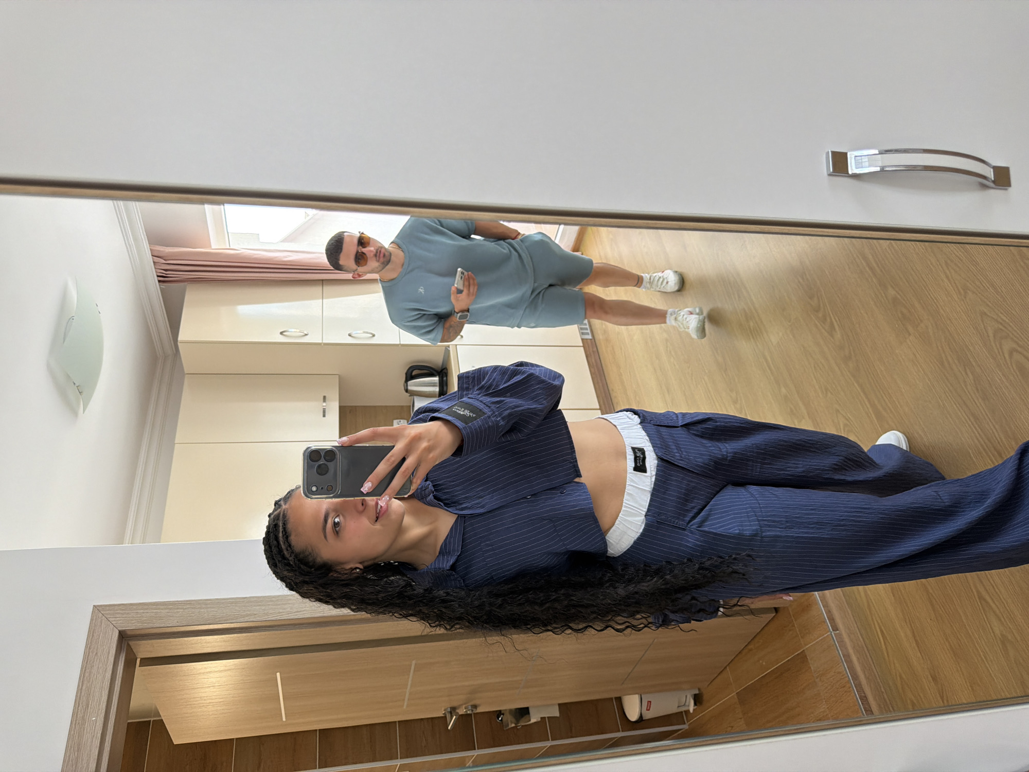The image shows a young woman with long, braided dark hair, wearing a navy blue pinstripe cropped shirt and matching wide-leg pants, taking a selfie in a mirror. Her pants have a white waistband. In the reflection behind her, a man wearing sunglasses, a light blue t-shirt, and matching shorts is visible, holding a smartphone. He is standing in what appears to be a hotel room with light-colored cabinets and a pink curtain. The room has wooden floors and a door on the left. The scene is consistent with a hotel room in Nesebar, Bulgaria, likely a modern apartment-style accommodation.