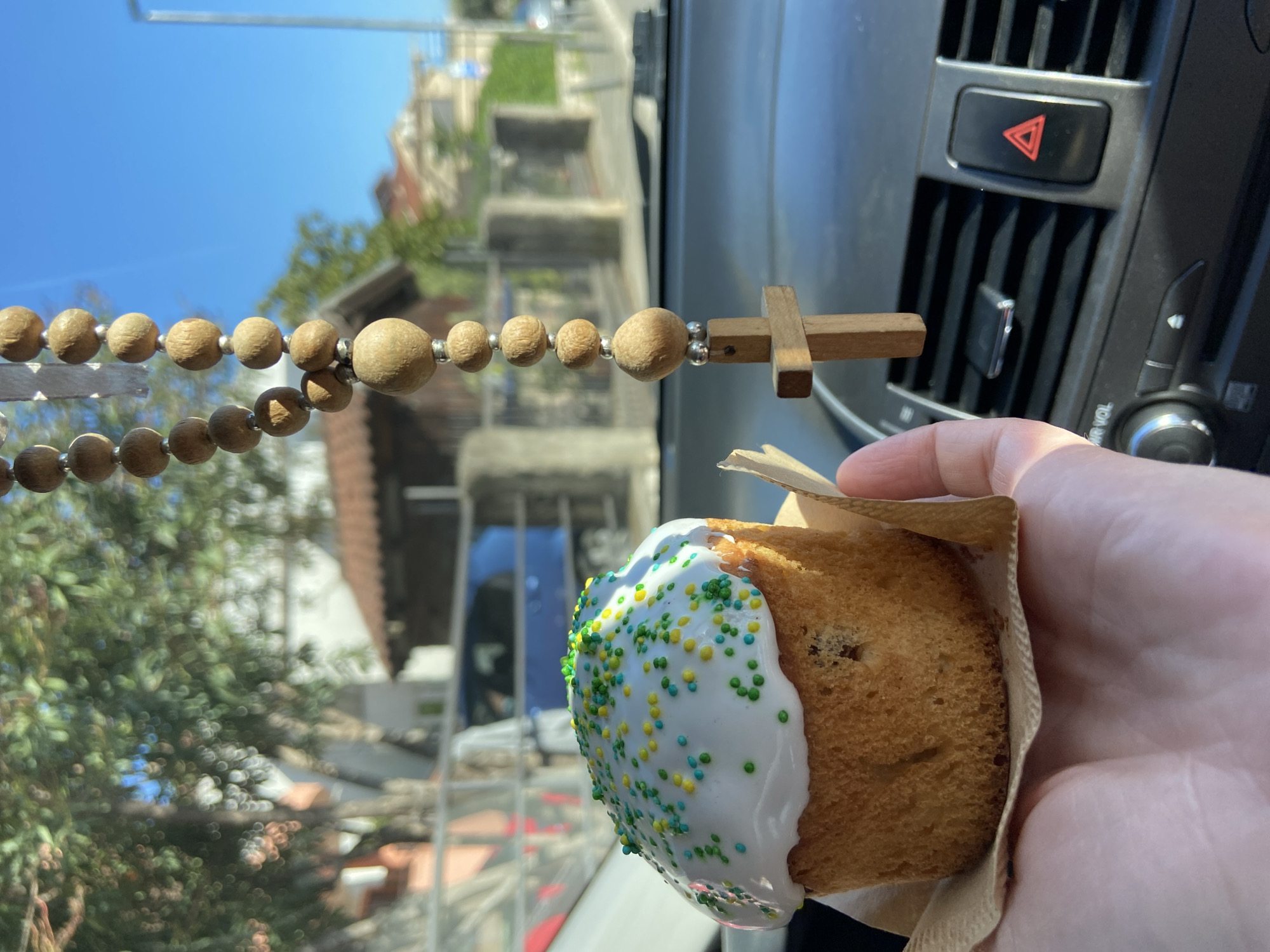 A photograph taken from the interior of a vehicle, possibly a car, features a hand holding a small cake or pastry with white icing and colorful sprinkles. The vehicle's dashboard and air vents are partially visible, along with the hazard light button. In the foreground, a string of wooden beads with a small wooden cross is suspended, possibly from the rearview mirror.
The background shows an out-of-focus cityscape, with buildings and greenery suggesting an urban environment with vegetation. The composition suggests the photograph may have been taken in or near Budva, Montenegro, given the architectural style and landscape visible through the vehicle's window.