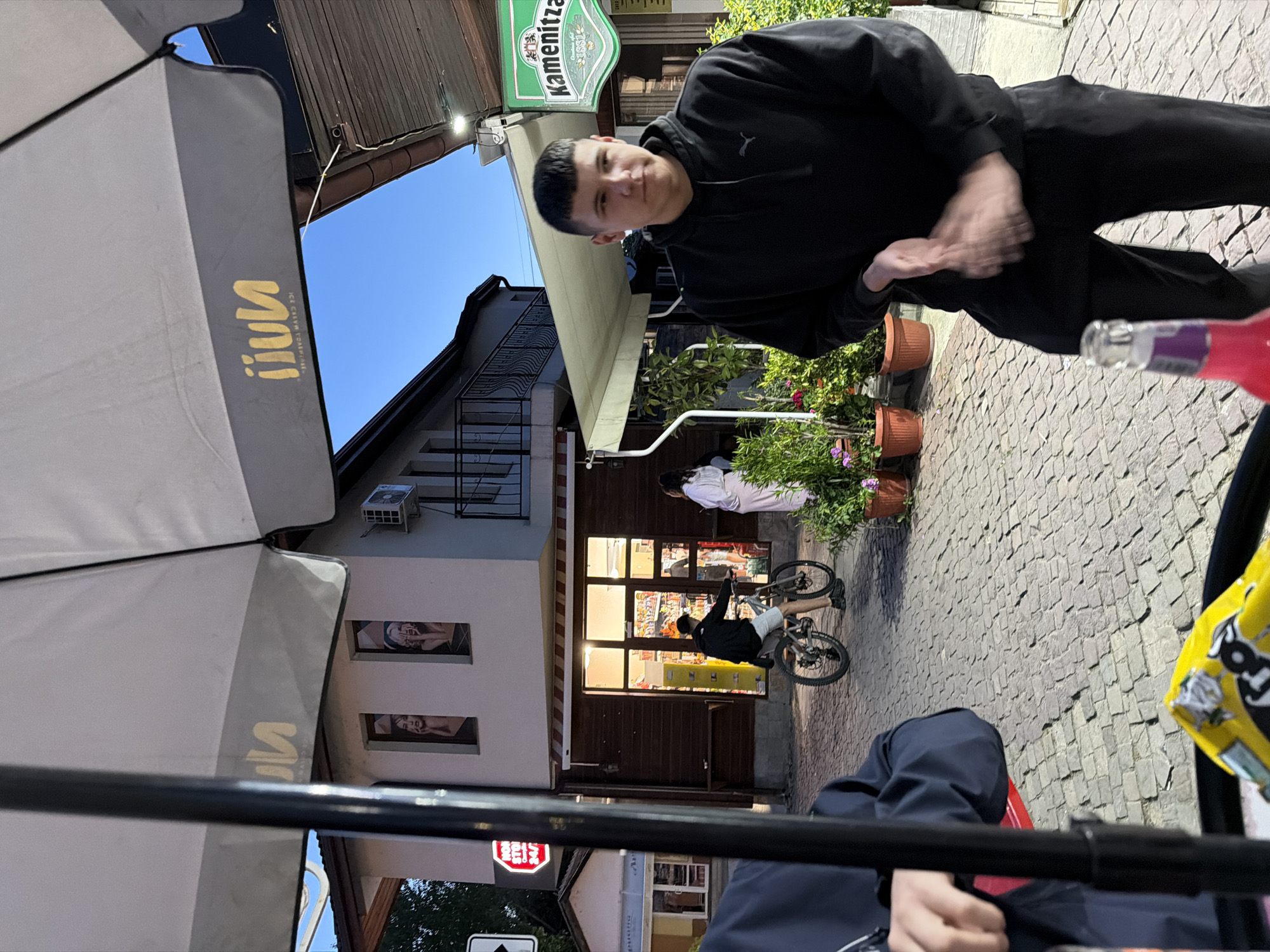 A street scene in Panagyurishte, Bulgaria features a young man standing near outdoor seating on a cobbled street, next to a restaurant or cafe with an awning. A child rides a bicycle in front of the building. Another person is standing in the doorway, possibly an employee. The "Kamenitza" sign suggests a local brewery. The "iluu" brand name is visible on an umbrella. The overall atmosphere suggests a casual, everyday scene in a European town. The angle is a bit skewed, and the focus is on the street scene. A person is visible in the foreground.