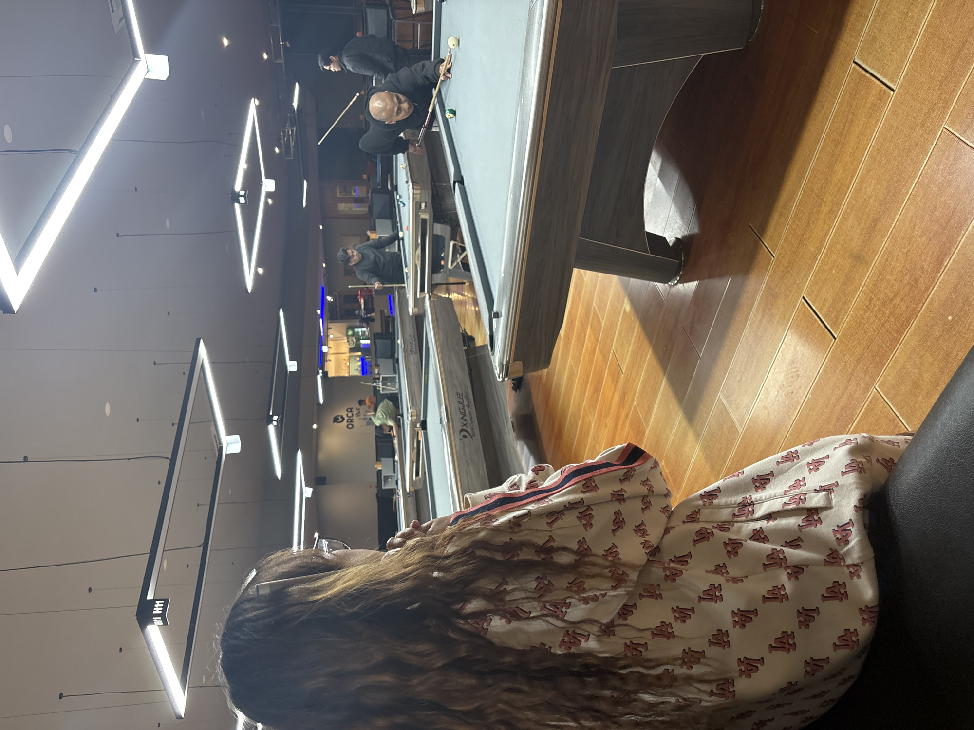 An indoor billiard hall in Kuta Selatan, Indonesia features several pool tables with overhead lighting. In the foreground, a woman with long, wavy brown hair and glasses is seated, facing away from the camera, wearing a light-colored jacket with a repeating "LA" logo pattern. In the background, three men are actively playing pool. One man, wearing a black hooded jacket, is in the process of taking a shot on a pool table in the mid-ground, with a pool cue in his hands. Another man, also in dark clothing, stands behind him, observing the game. A third man, in a green shirt, is visible at another pool table further in the background, also playing. The environment is a well-lit commercial space with polished wooden floors.