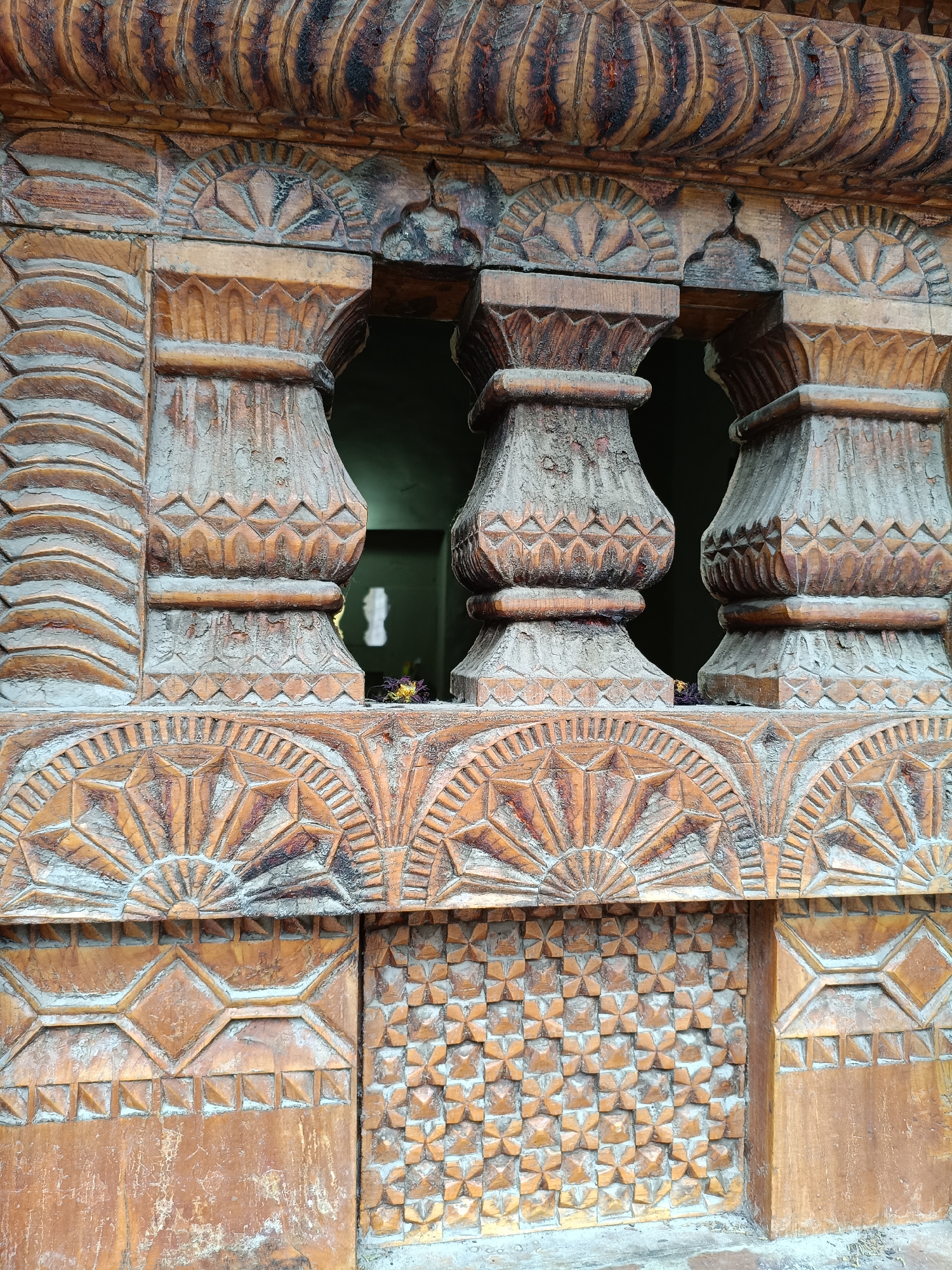 The image displays a close-up of intricately carved wooden lattice work, likely part of a window or architectural feature. The wood is a warm, reddish-brown color, with areas of darker staining and a visible texture that suggests age and weathering. The carving is detailed and geometric, featuring patterns that resemble sunbursts, diamond shapes, and repeating floral or star-like motifs.
The lattice has a series of carved pillars that divide the openings. These pillars have a bulbous, turned shape, adorned with faceted patterns. Above the pillars, a more ornate carved band continues the decorative theme, with semi-circular, fan-like designs. The overall impression is one of traditional craftsmanship.
Some of the carved surfaces show a fine layer of dust or residue, indicating the piece is either outdoors or has not been recently cleaned. In the dark openings between the pillars, there are faint hints of color, possibly from dried flowers or other small objects that have been placed there. The lighting is even, suggesting it is daytime, but there are no strong shadows to indicate a specific time. The context provided places this scene in Vashisht, India, which is known for its ancient temples and traditional architecture. No text is visible in the image.