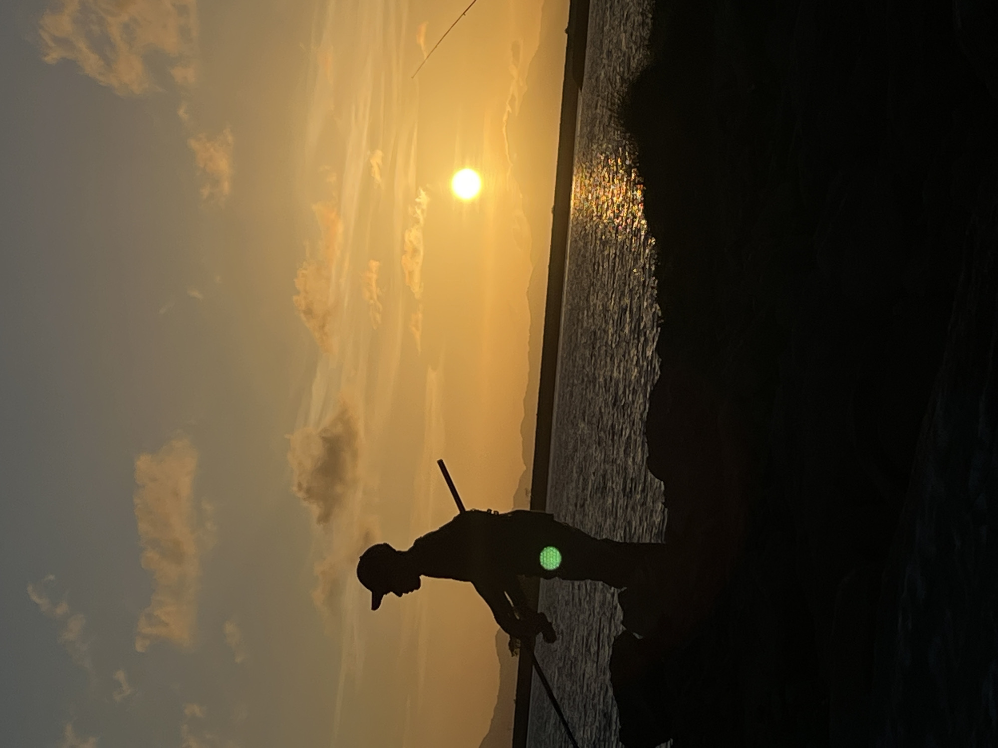 This is a silhouetted image of a person fishing at sunset on the coast of Banda Aceh, Indonesia. The sun is low on the horizon, casting a warm orange glow across the sky and water. The person is standing on a rocky shore, holding a fishing rod. The water is calm, with gentle waves lapping at the shore. In the distance, mountains can be seen silhouetted against the sunset. The overall mood of the image is peaceful and serene.
