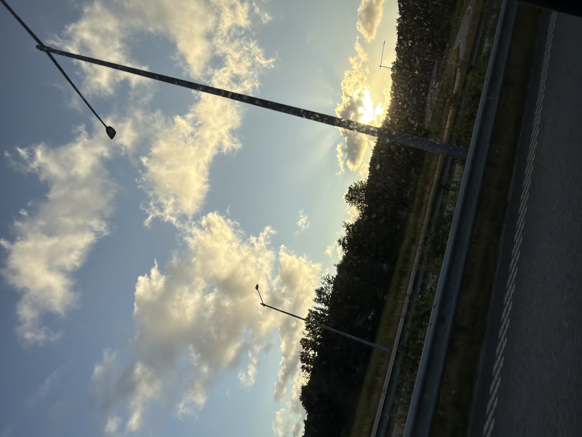 The image depicts a sky with scattered clouds, illuminated by a setting or rising sun. Sunbeams are visible radiating from behind the clouds. In the foreground, a dark road with white lane markings is visible, separated from a grassy verge by a metal guardrail. A tall street lamp with a curved arm and a dark fixture stands prominently on the right side of the frame, silhouetted against the sky. Another street lamp is visible further in the background on the left. A dense line of dark trees forms a backdrop behind the grassy verge. The overall impression is of a roadside scene during twilight. The location is Ödsmål, Sweden.