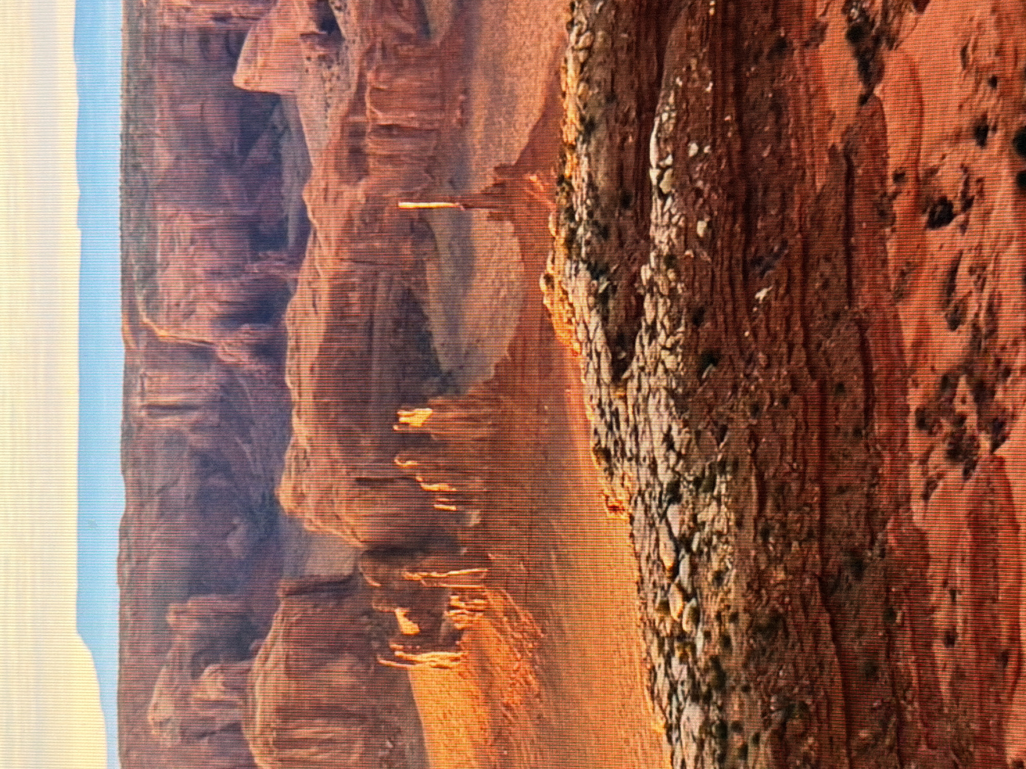 This is a wide-angle, landscape photograph of a desert canyon. The image is dominated by warm, reddish-orange hues from the sandstone formations. In the foreground, a layered rock formation is visible, with smaller stones and sparse vegetation. In the mid-ground, several tall, slender rock spires rise from the canyon floor, illuminated by a warm, golden light that suggests either sunrise or sunset. Further back, the canyon walls rise in layered formations, with deep shadows creating a sense of depth. The background shows a distant, flat-topped mountain range under a pale blue sky. There are no people or animals clearly visible, though a small, reddish-orange blur in the mid-ground might indicate a distant figure or animal. The scene depicts a natural, remote, and arid environment. There is no indication that this location is in Kyiv, Ukraine.