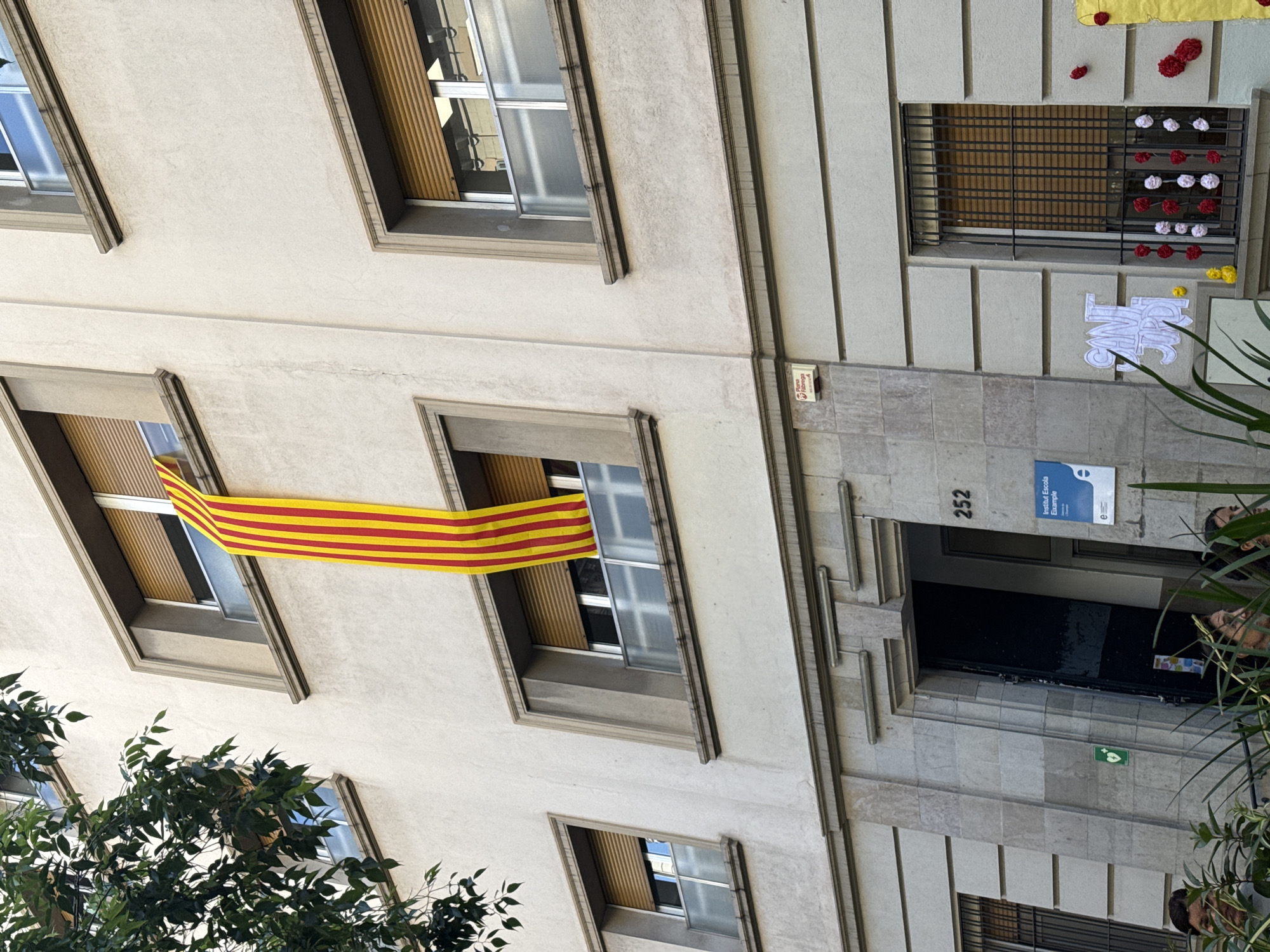 This vertical shot captures a multi-story building facade in Barcelona, Spain, likely a public institution, during daylight hours. The building features light-colored walls with stone accents and multiple windows, some fitted with wooden blinds.
A prominent yellow and red striped flag, consistent with the Catalan Senyera, is draped from a window on the lower-left side of the building. On the upper-right side, a barred window is adorned with decorative paper flowers in red, white, and yellow, and a white paper cut-out decoration is placed beneath it.
The main entrance, situated on the right side of the image, is a dark, recessed doorway. Above this entrance, the building number "252" is clearly visible on a stone pillar. A sign next to the entrance identifies the location as "Institut Escola Eixample," with an accompanying logo. Further details near the entrance include a red box with partial text "Fiarega" and a small green square sign with a white symbol, possibly indicating an emergency point or first aid.
Green foliage from trees and plants is present in the foreground on both the left and right sides, partially obscuring the lower windows and a few individuals near the building's entrance. The visible decorations and the school identification suggest an active public building.