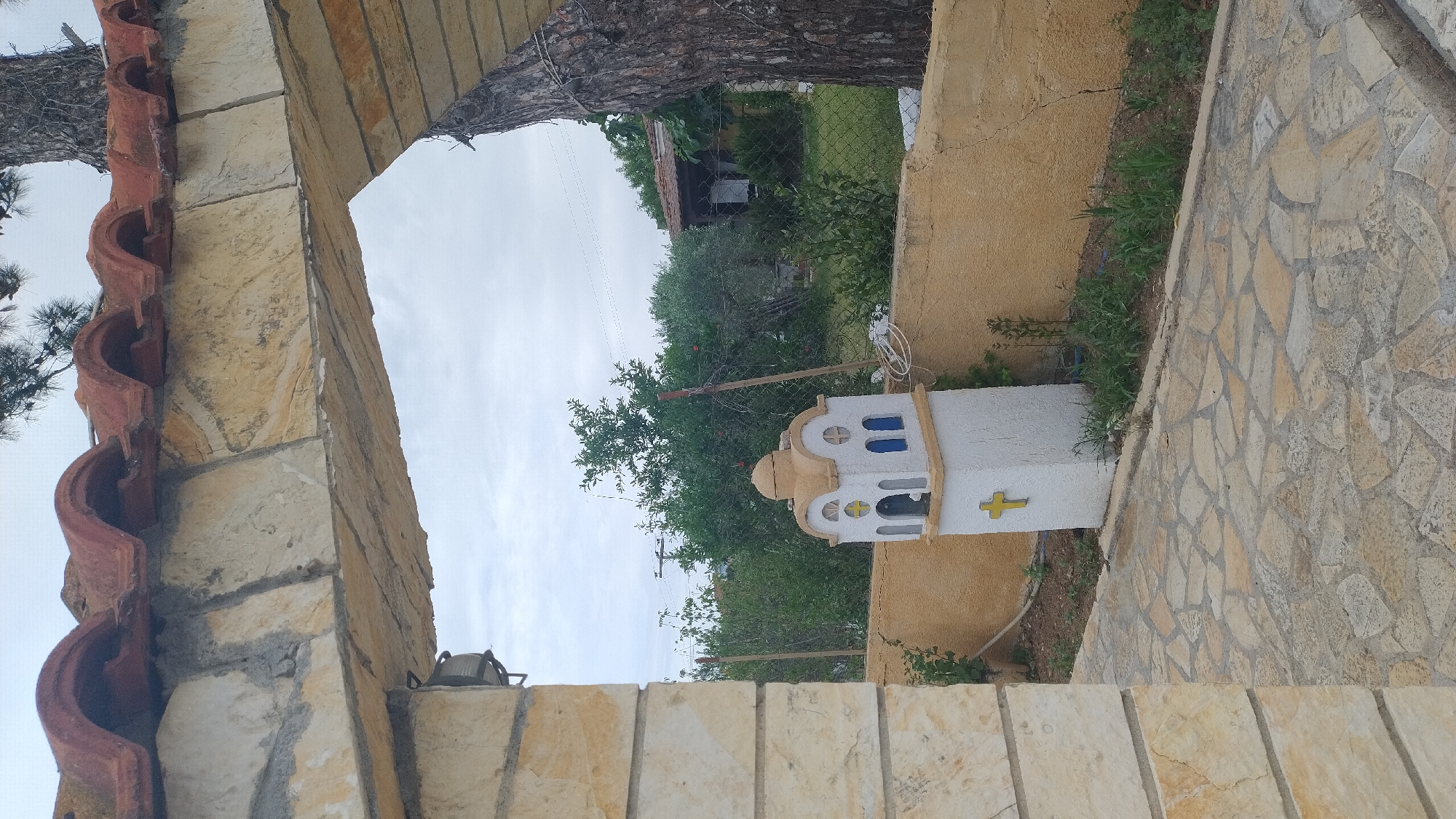 The image shows an outdoor scene viewed through a stone archway with a tiled roof. In the foreground, a pathway made of irregularly shaped stones leads towards the interior of a courtyard. To the left, a stone pillar supports the archway. In the middle ground, a small, white, steeple-like structure with a dome is visible, featuring blue arched windows and a yellow cross on its side. This structure is situated in front of a yellow stucco wall. Behind the wall, a fence separates the courtyard from a garden area with lush green trees and vegetation. A rustic building with a tiled roof is partially visible in the background. The sky is overcast, and a large tree trunk is prominent on the right side of the image, leaning into the archway. The scene appears to be in Νέα Κερασιά, Greece, likely a residential or religious site given the presence of the miniature church-like structure. There are no people or animals visible in the image.