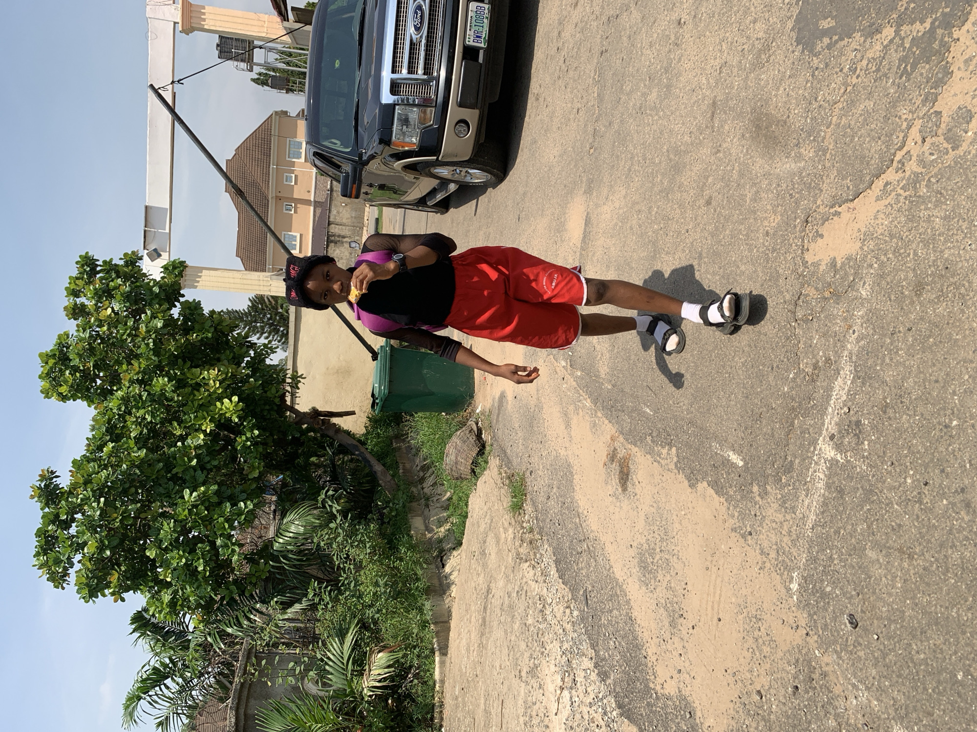 A young person walks on a paved street in Abuja, Nigeria, holding a piece of food up to their mouth. They are wearing a black shirt, red shorts, white socks, and black sandals. A purple backpack is slung over their shoulders. To the right of the person is a black Ford pickup truck. In the background, there is a building, a large green trash bin, and lush green trees. The sky is clear and blue.