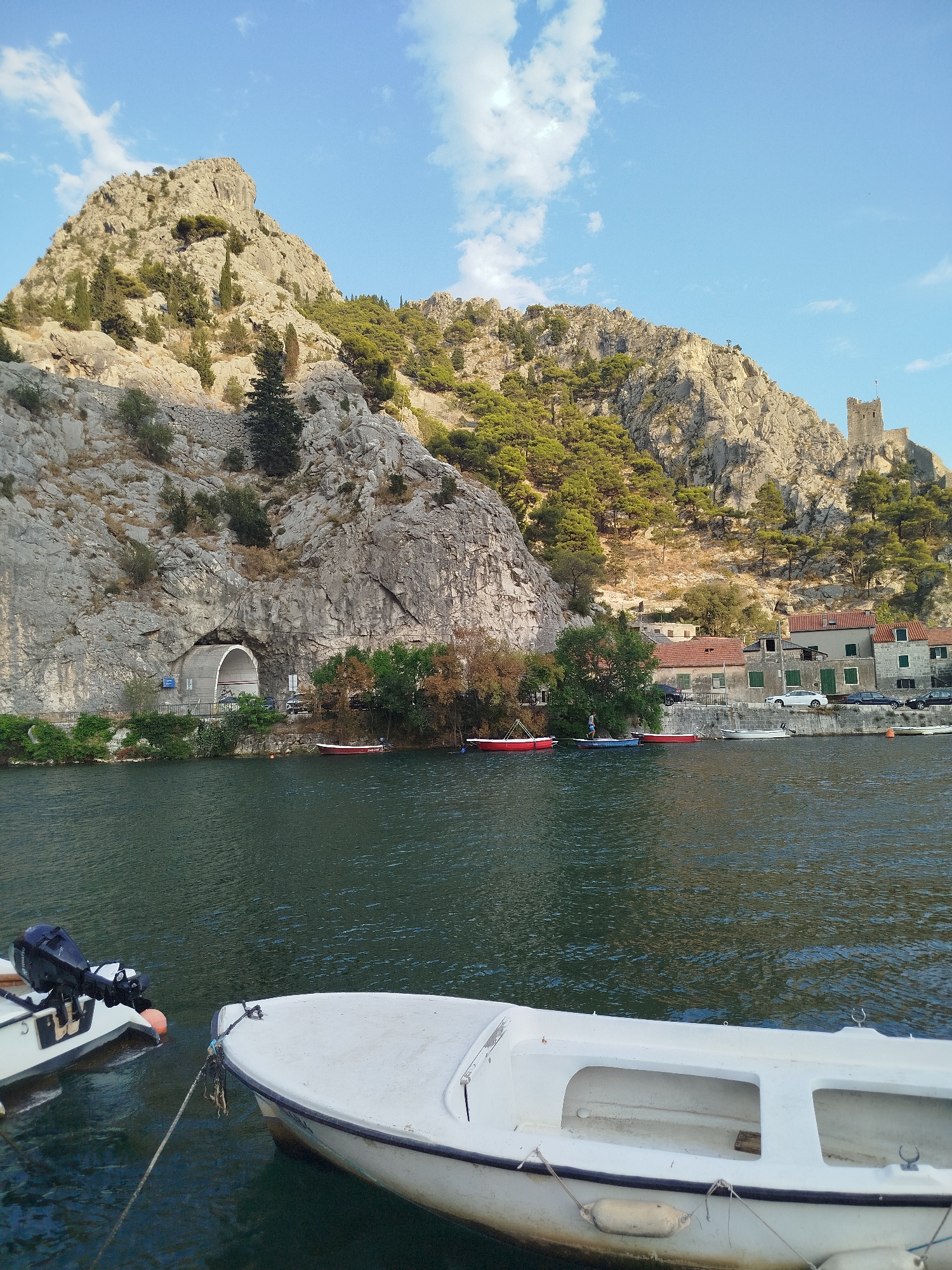 This is a landscape photograph taken from a body of water, likely a river or inlet, looking towards a rocky mountainside and a small town. In the foreground, the bow of a white boat is visible, tied to a mooring line. Another boat, partially visible to the left, has an outboard motor.
The midground features a calm body of dark water. Along the shoreline, several small, colorful boats are docked or moored. Behind the boats, a row of buildings with red-tiled roofs lines the waterfront. These buildings appear to be part of a residential or commercial area. Behind the buildings, the steep, rocky mountainside rises dramatically. A prominent feature of the mountainside is a large tunnel entrance carved into the rock. Trees, predominantly pine trees, are scattered across the slopes, with some denser vegetation near the base of the mountain. A stone structure resembling a castle or fortress is visible at the very top of the mountain on the right side of the image.
The sky above is a clear blue with scattered white clouds. The lighting suggests it is either early morning or late afternoon, with sunlight illuminating the mountainside. The scene depicts the town of Omiš, Croatia, specifically the area where the Cetina River meets the Adriatic Sea, known for its dramatic karst landscape and historical fortresses.