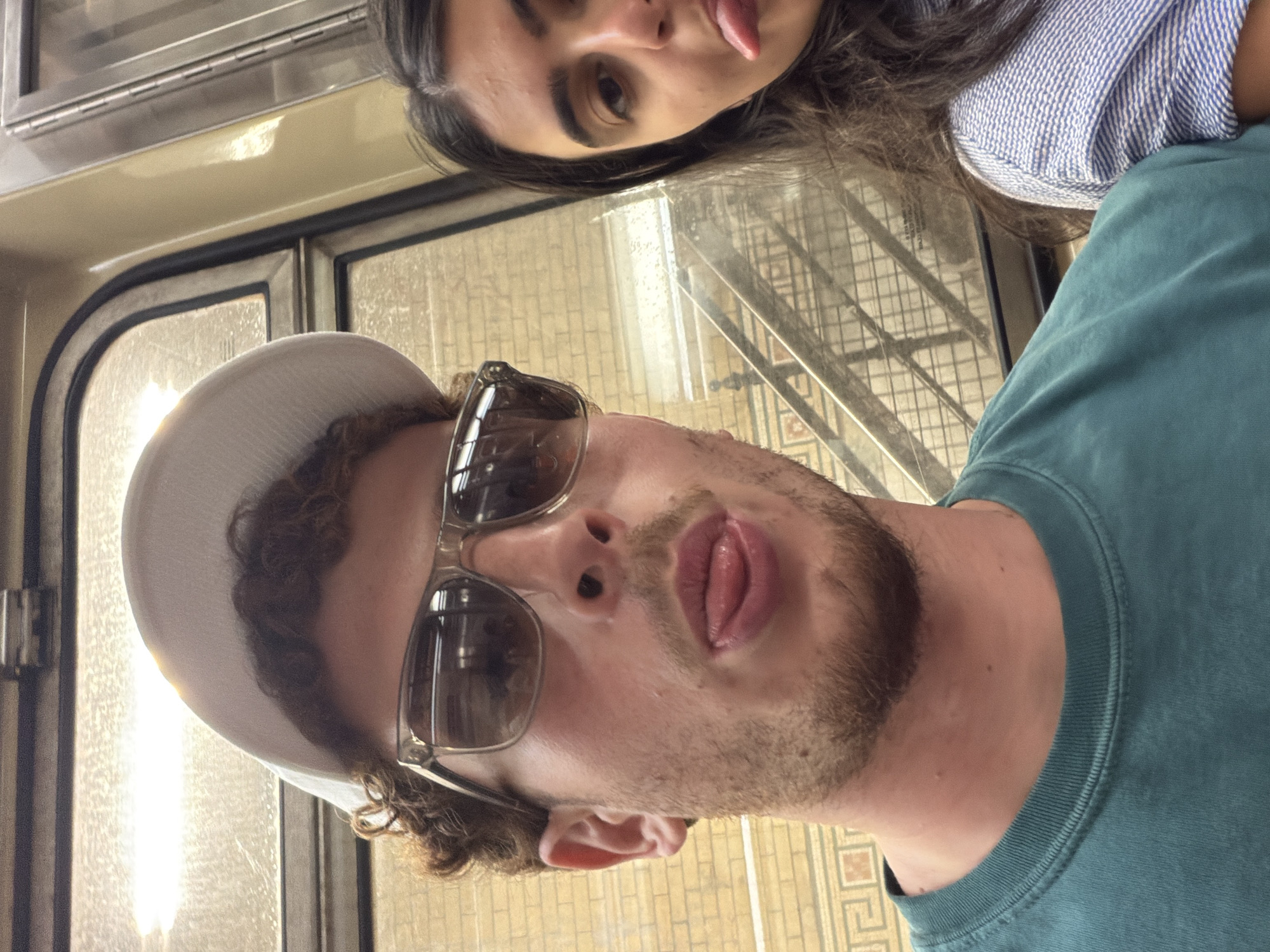 Two people pose for a selfie on a New York City Subway train. The man in the foreground wears sunglasses and a hat, sticking his tongue out. The woman in the background also sticks her tongue out. The train car's interior, including windows and tiled walls, is visible.
